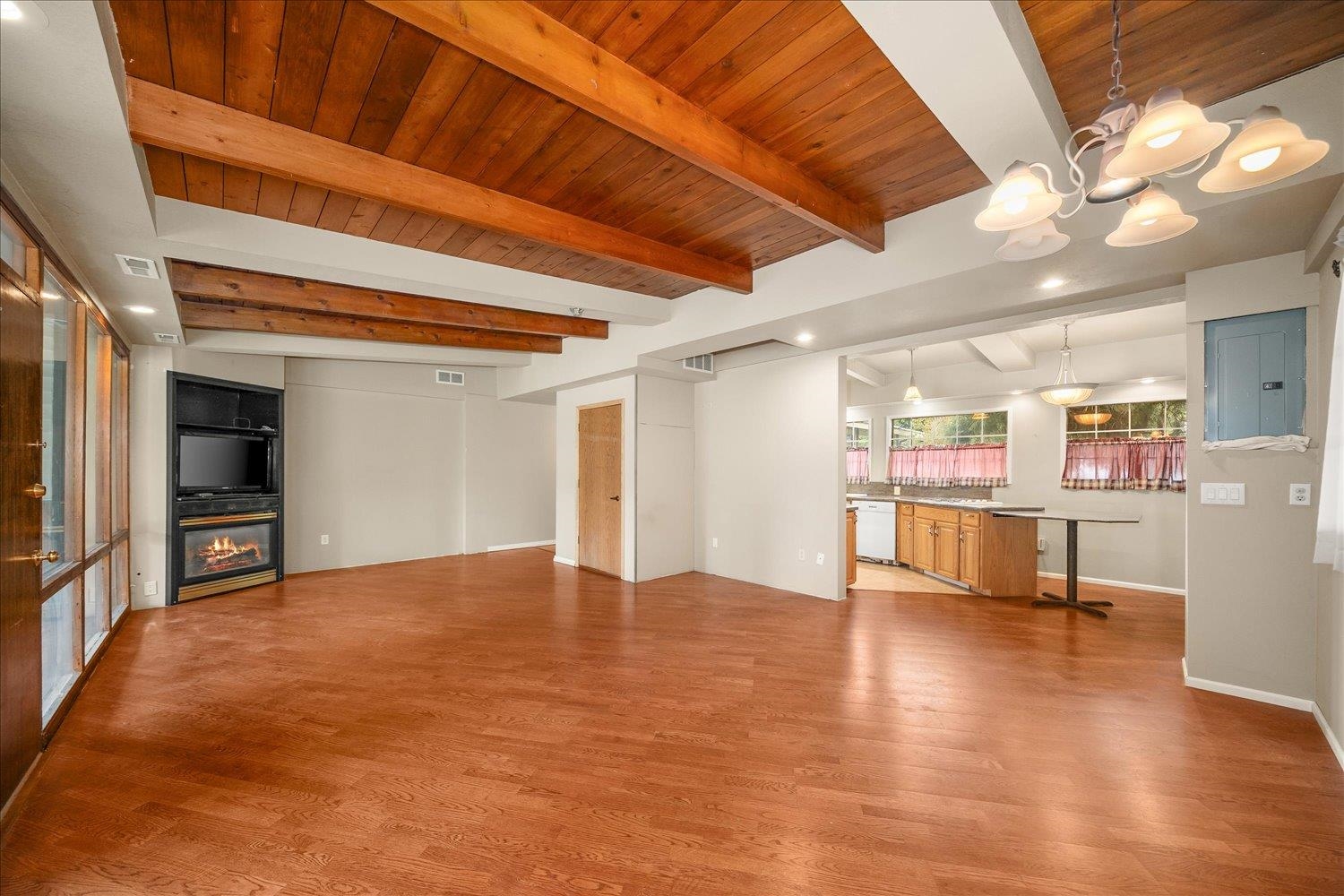 Open living room with hardwood floors, exposed wood ceiling beams, and a built-in fireplace beside the kitchen.