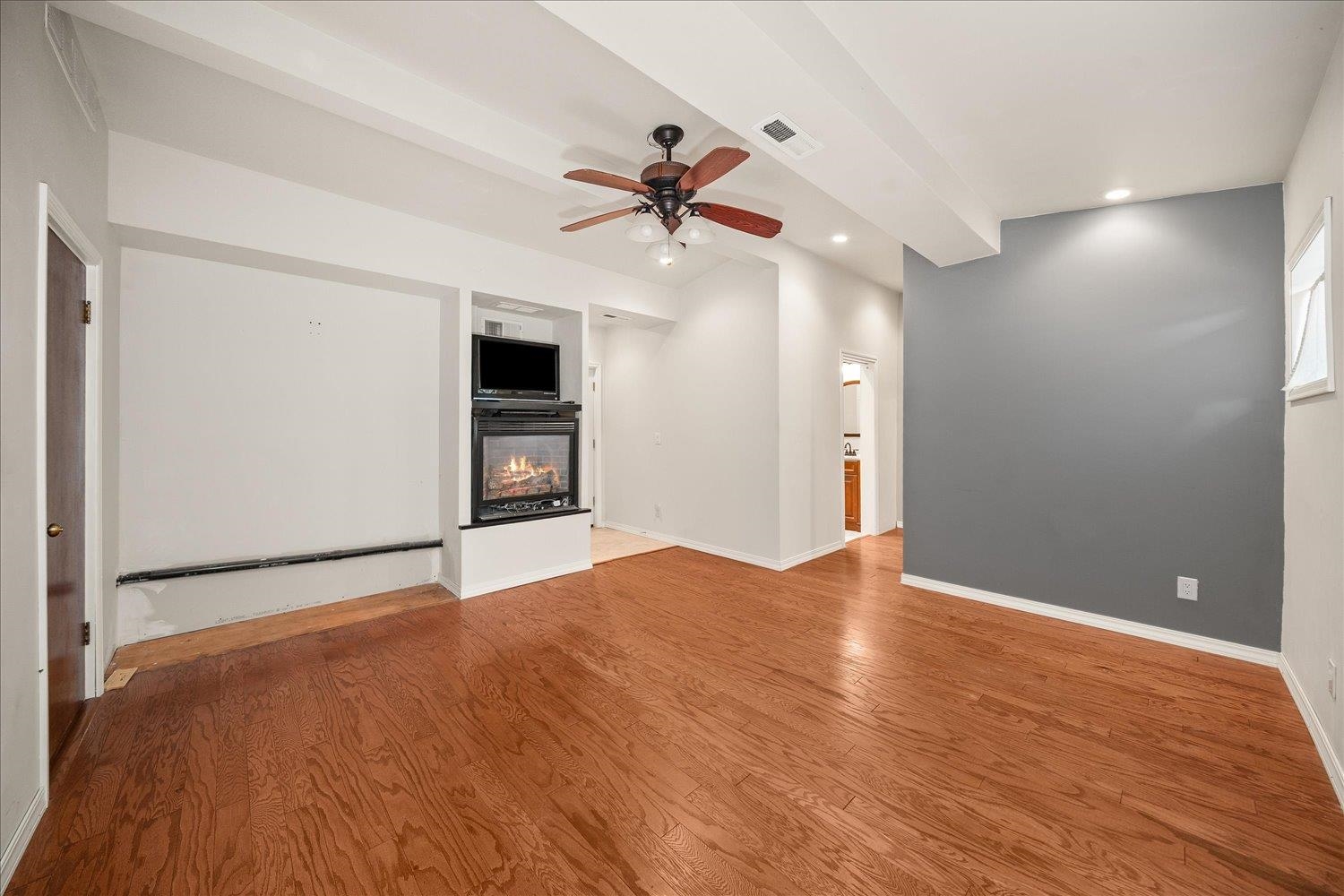 Empty living room with hardwood floors, a gray accent wall, and built-in fireplace beneath a beamed ceiling.