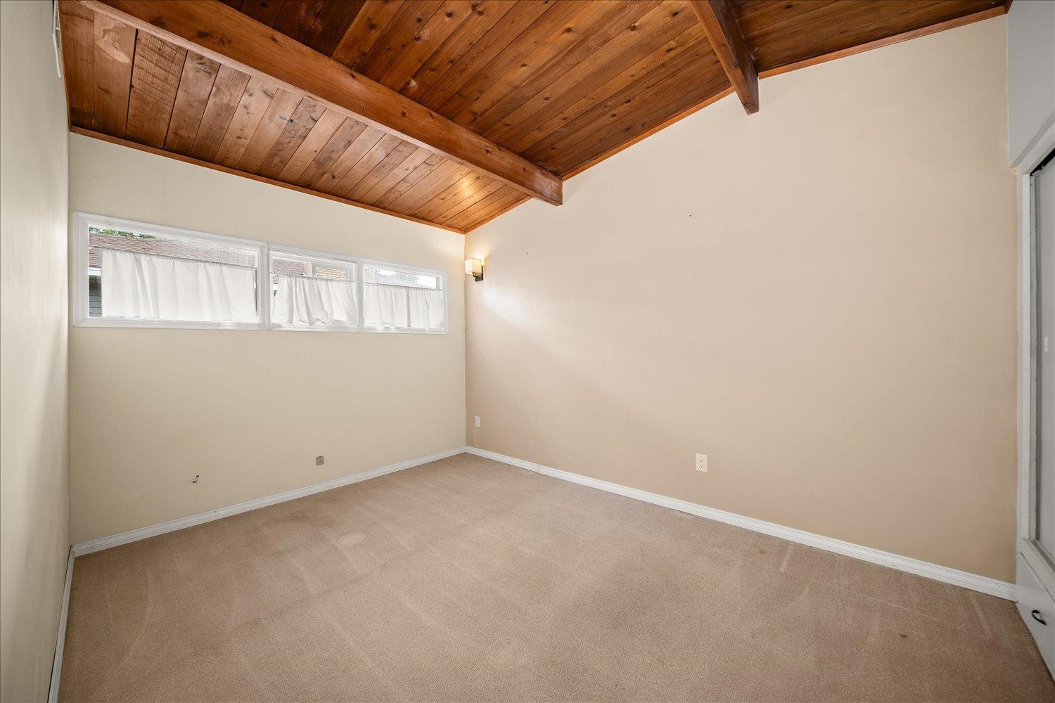 Empty carpeted room with a vaulted wood ceiling and exposed beams.