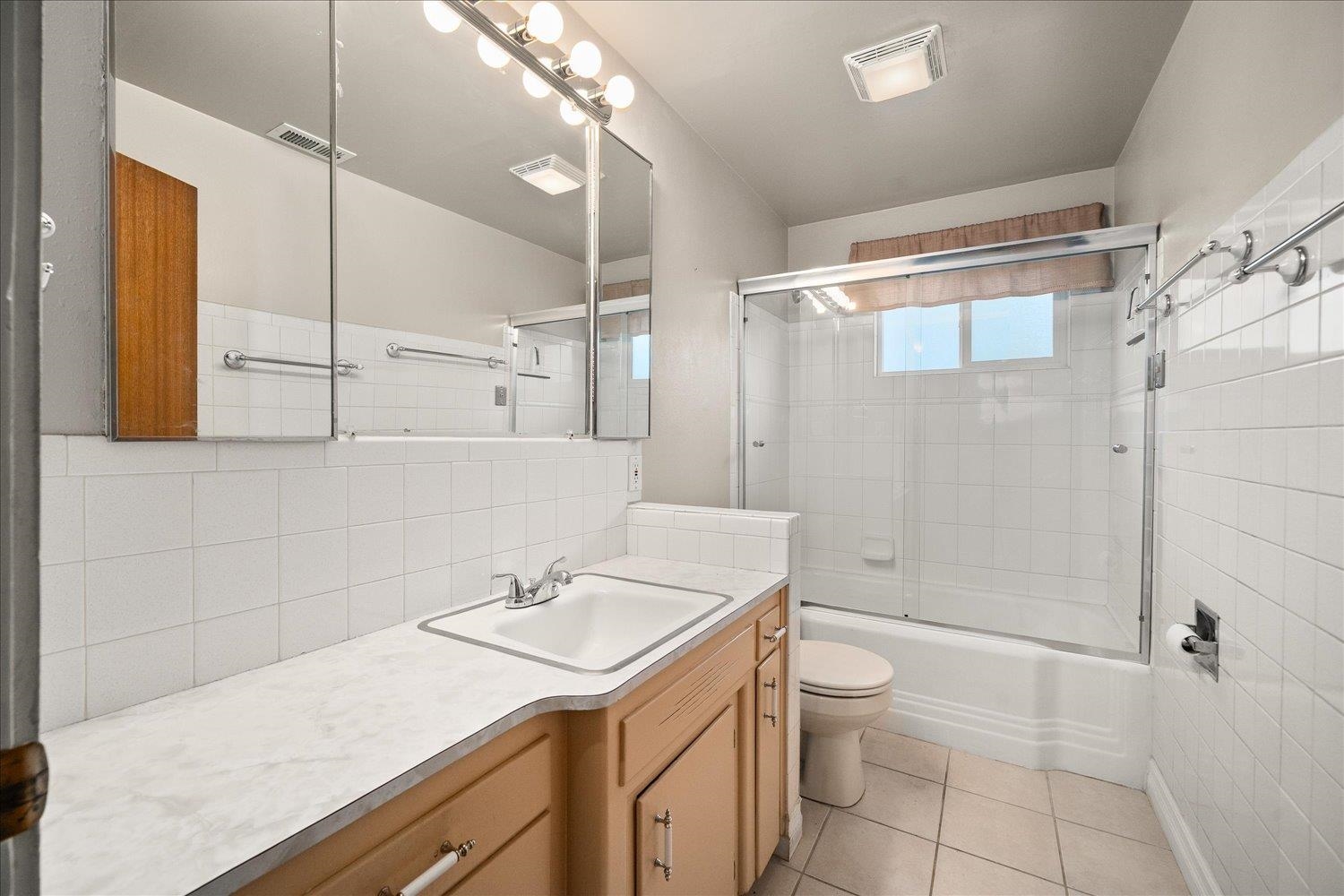 Bathroom with a long wood vanity, mirrored cabinet, and white-tiled tub-shower enclosure.