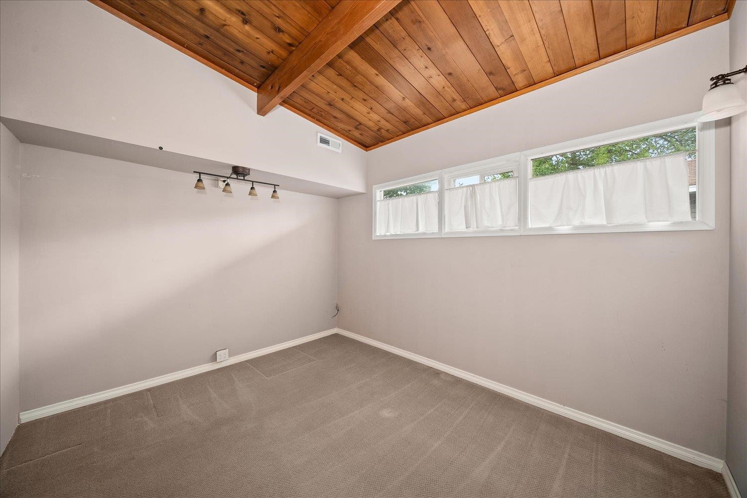 Empty carpeted room with a vaulted wood ceiling, exposed beam, and curtained windows.