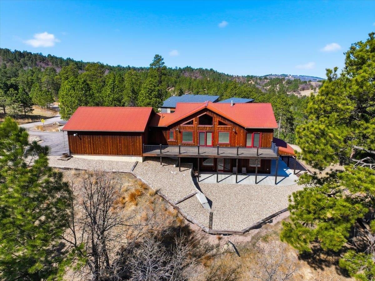 Wood-sided home with red roofs and a raised deck, surrounded by trees and wooded hills.