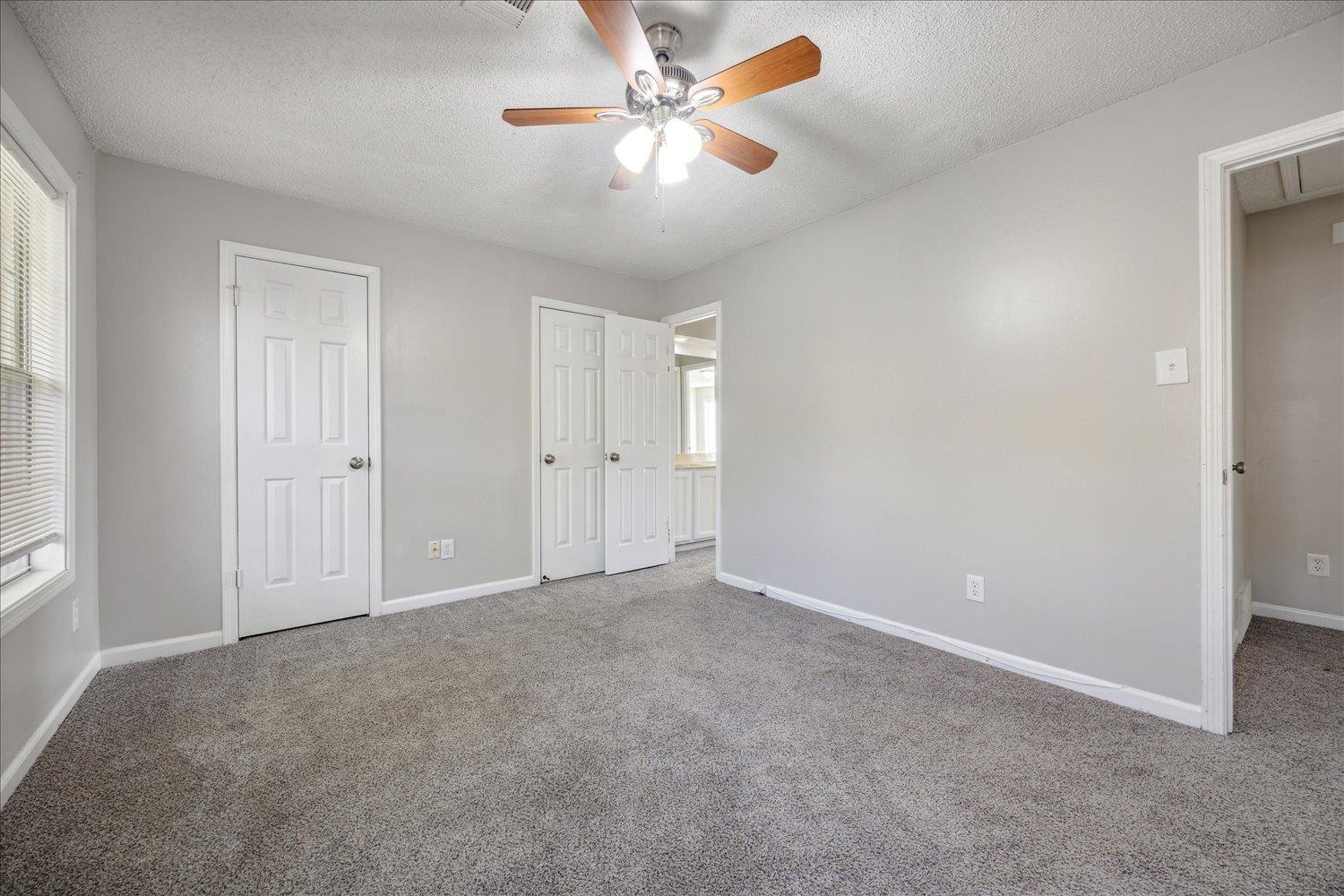 Empty bedroom with gray carpet, a wooden-blade ceiling fan, and open closet doors.