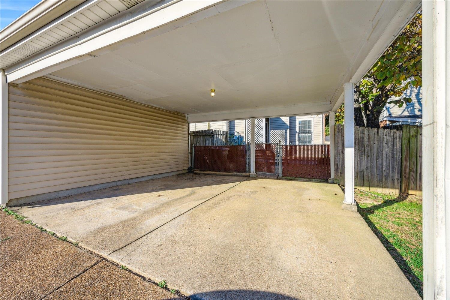Covered carport with a wide concrete driveway and red metal gate at the far end.