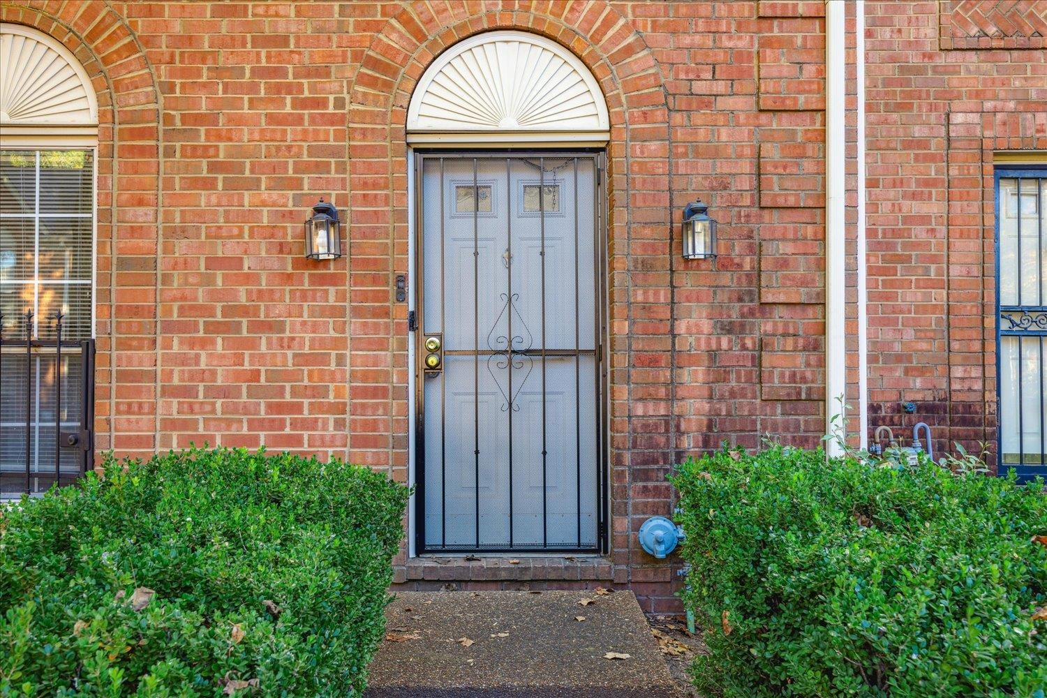 Brick home entrance with an arched doorway, metal security door, and trimmed shrubs.
