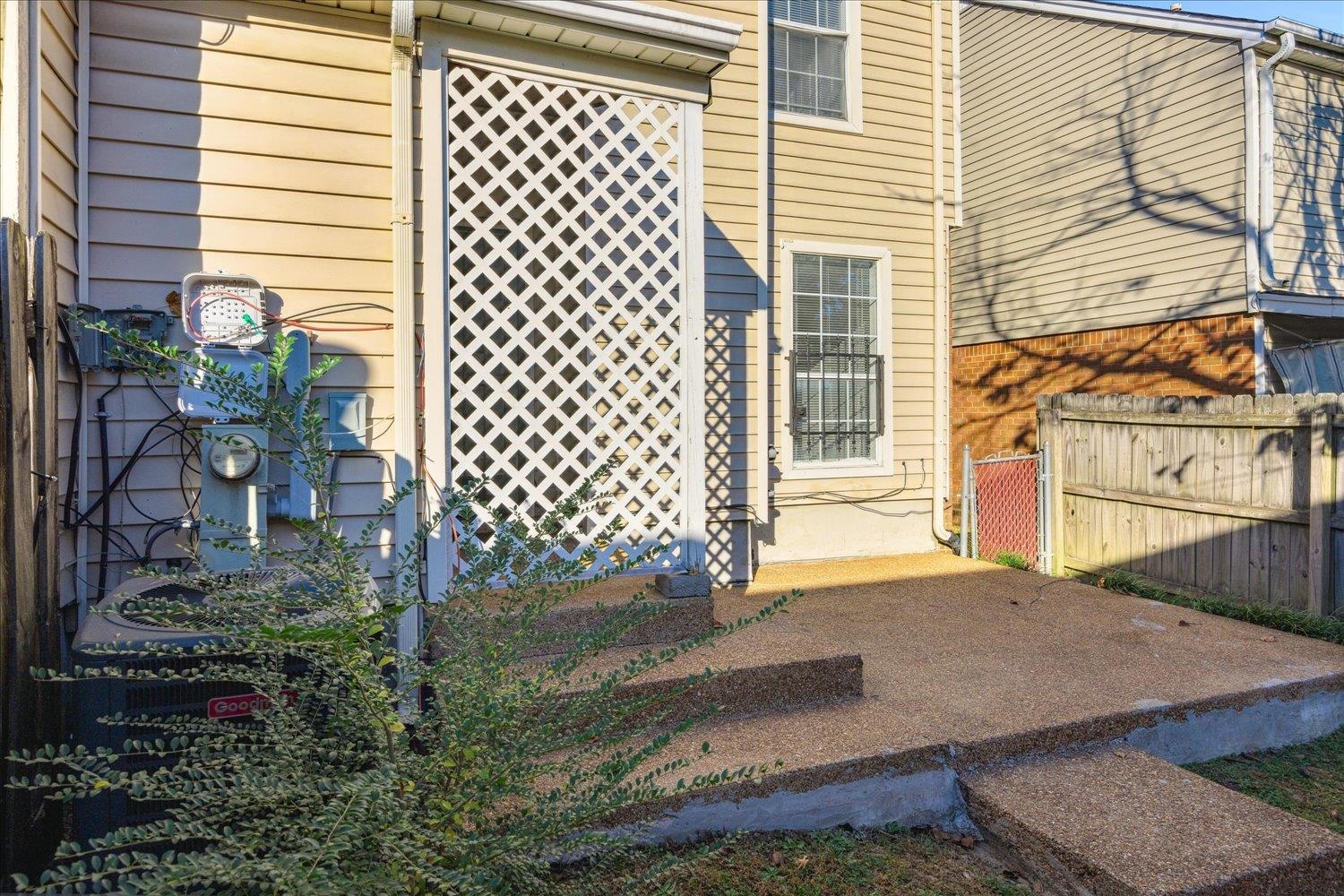 Rear side of a tan-sided home with a white lattice screen beside a concrete walkway and wooden fence.