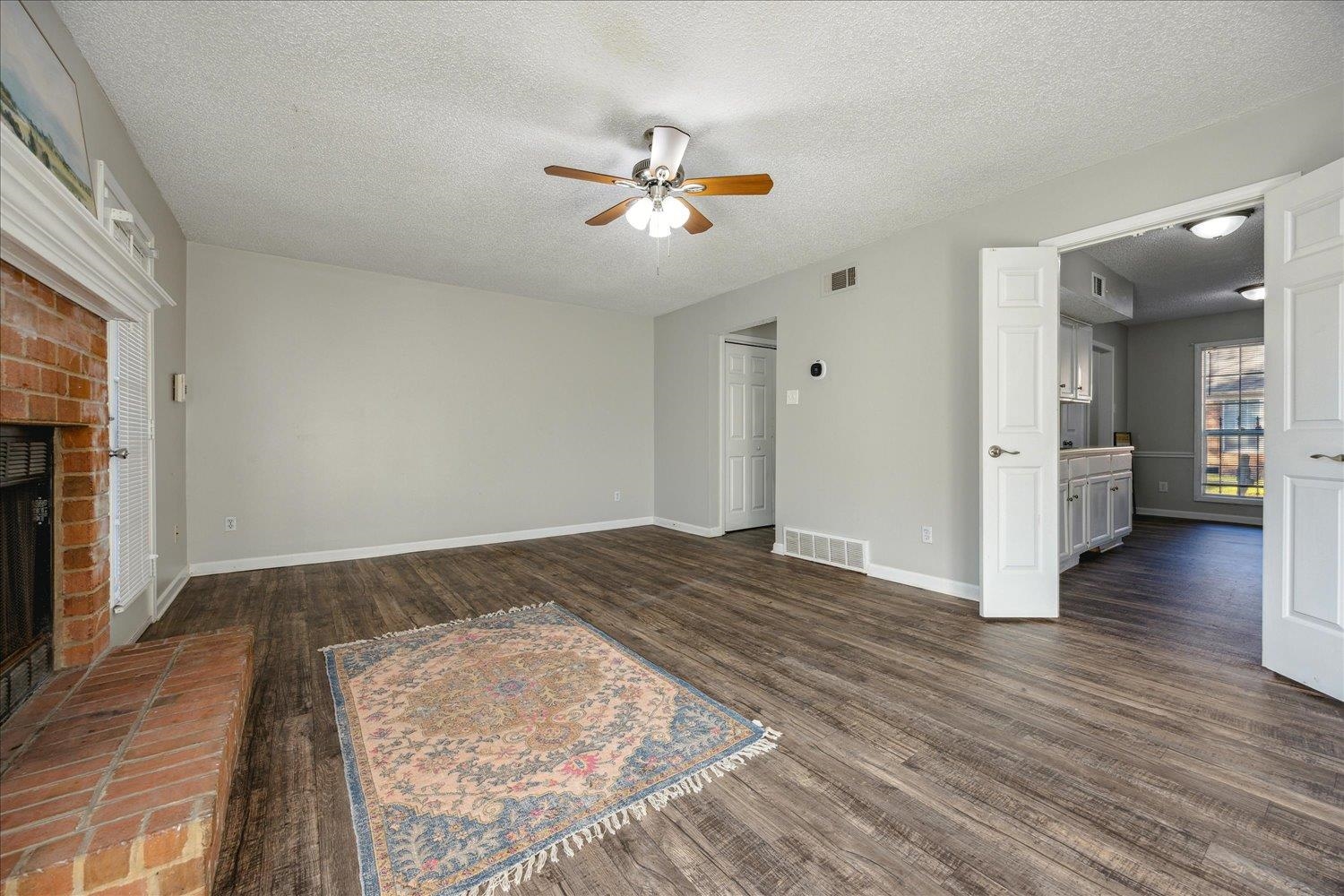 Living room with a brick fireplace, dark flooring, patterned rug, and open doors to the kitchen.