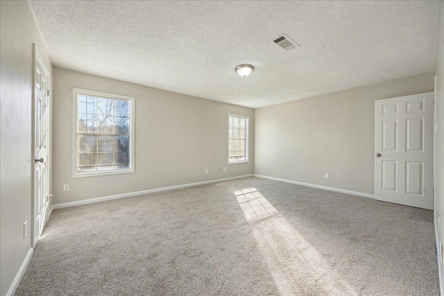 Empty carpeted bedroom with two windows and sunlight across the floor.