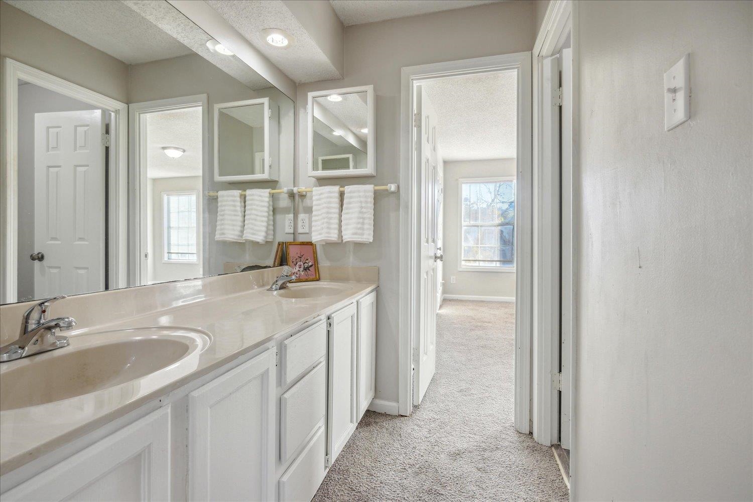 Bathroom with a long double-sink vanity, paneled cabinets, and mirrors overlooking a carpeted bedroom.