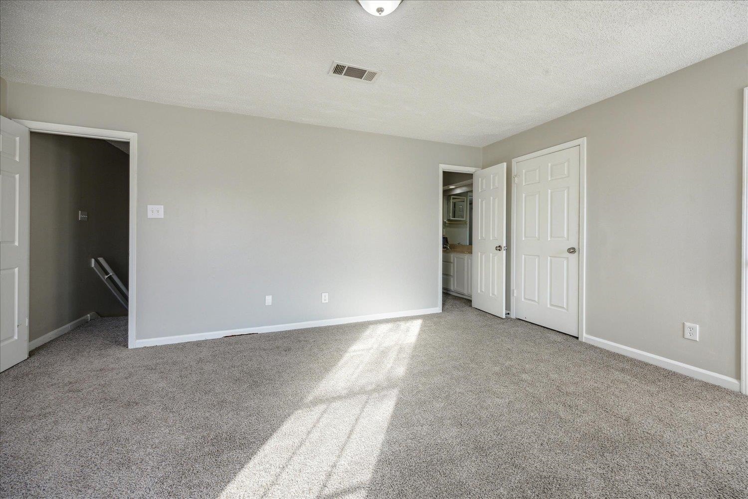 Empty bedroom with gray carpet and sunlight across the floor, opening to a bathroom.