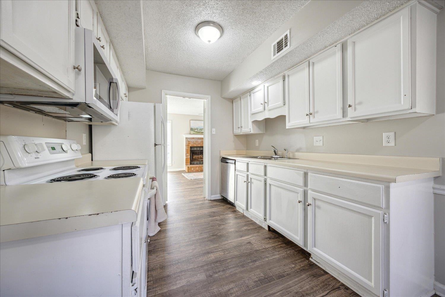 Kitchen with white cabinets and long counters, dark wood-look flooring, and a view toward a brick fireplace.