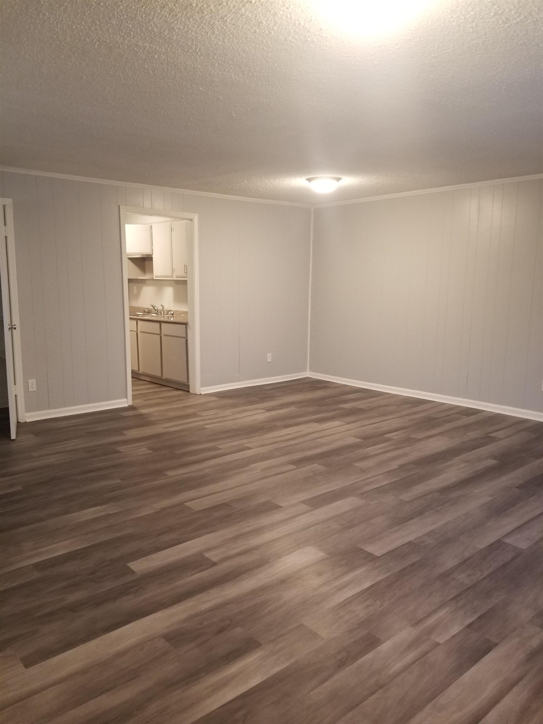 Empty room with gray paneled walls and wood-look flooring, opening to a compact kitchen.