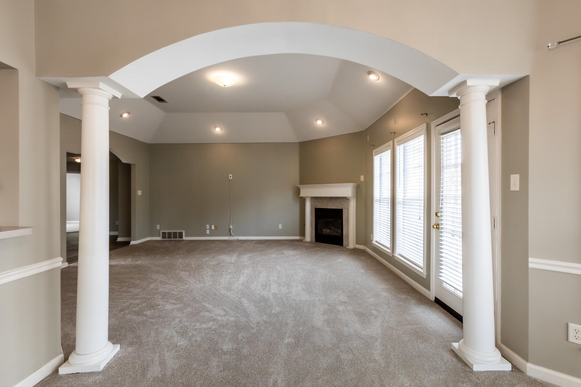 Vacant living room with a white fireplace, tall blinded windows, and an arched ceiling supported by columns.