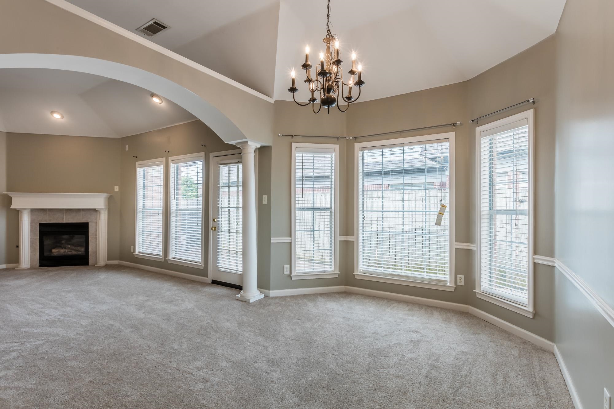 Empty living room with a fireplace, vaulted ceiling, chandelier, and a wall of tall windows.