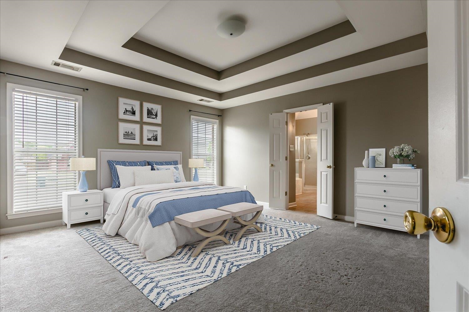 Staged bedroom with a layered tray ceiling, blue-and-white bedding, and an open bathroom doorway.