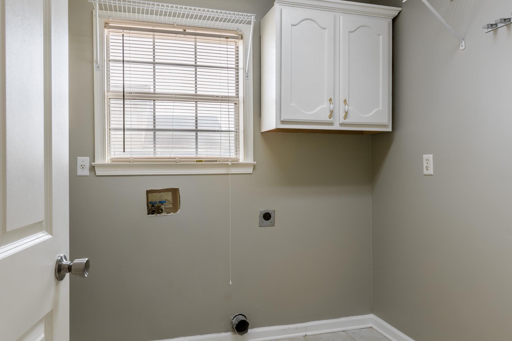 Empty laundry room with upper cabinet, window, exposed washer hookups, and dryer vent.