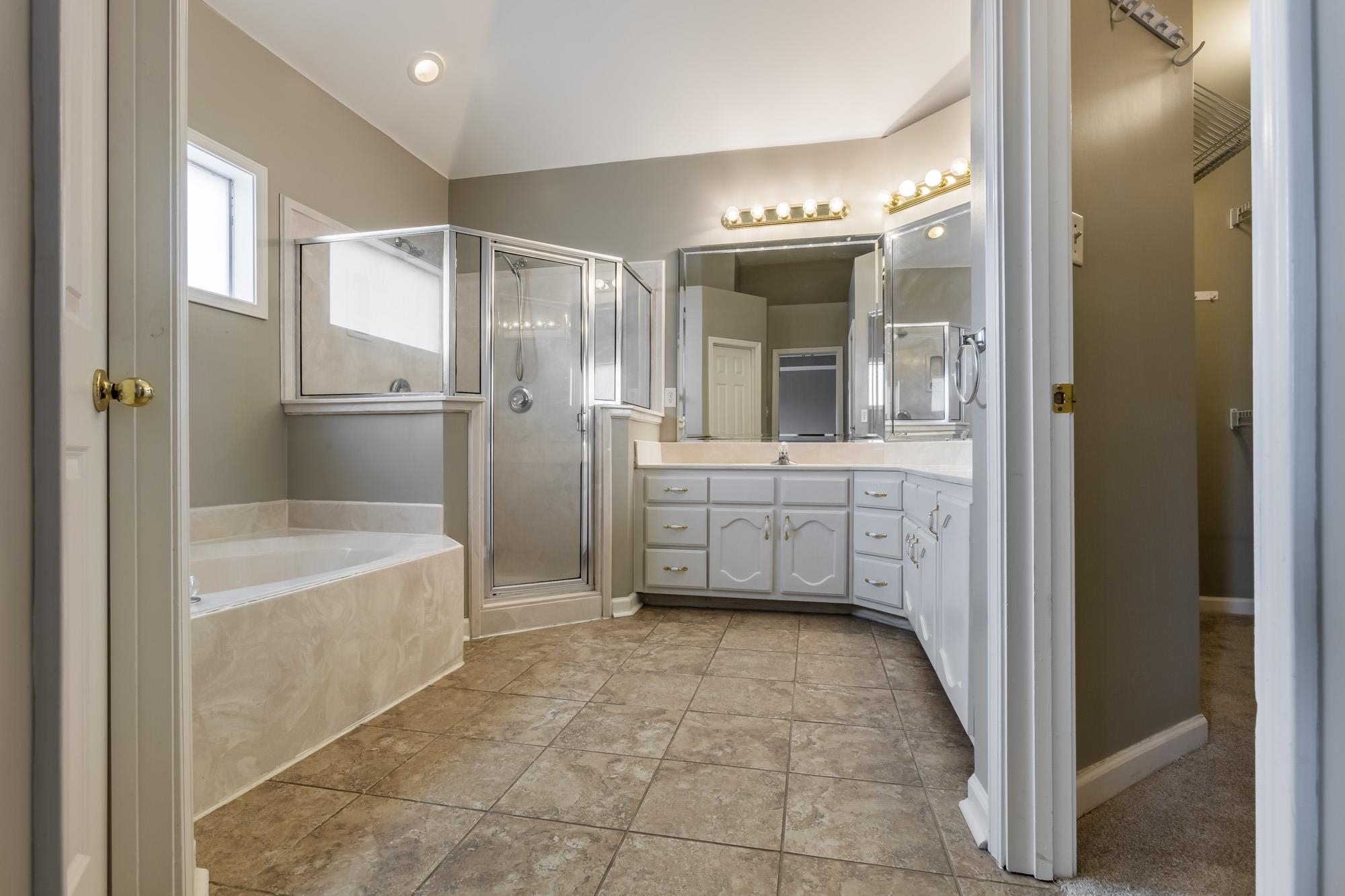 Bathroom with a corner soaking tub, glass shower, long white vanity, and brown tile floor.