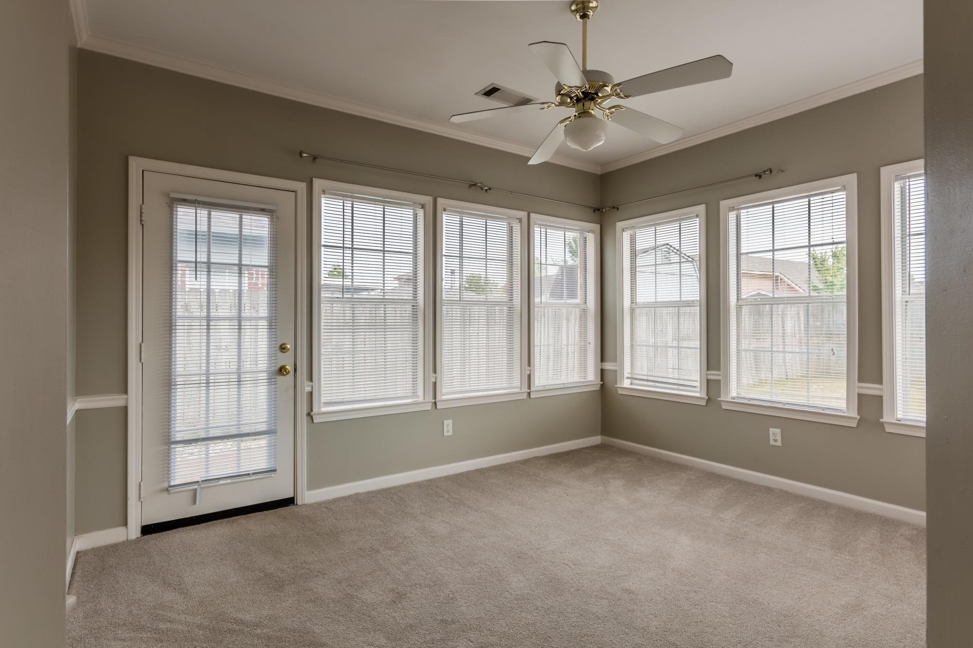 Empty carpeted sunroom with a ceiling fan, glass door, and rows of windows with blinds.