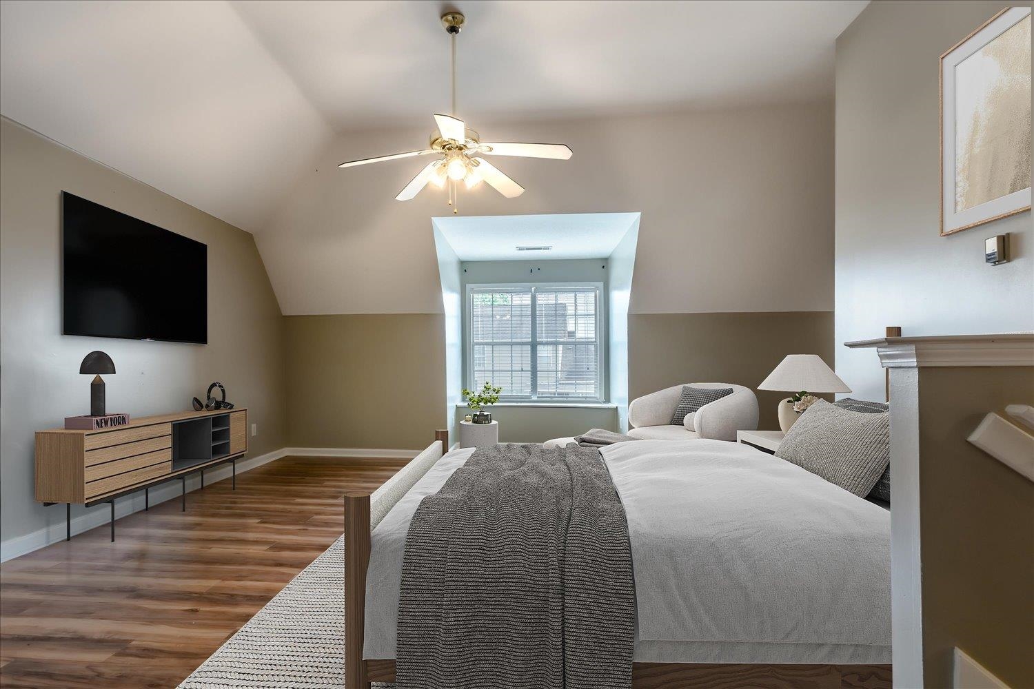 Furnished bedroom with vaulted ceiling, centered window, wall-mounted television, and gray-and-white bedding.