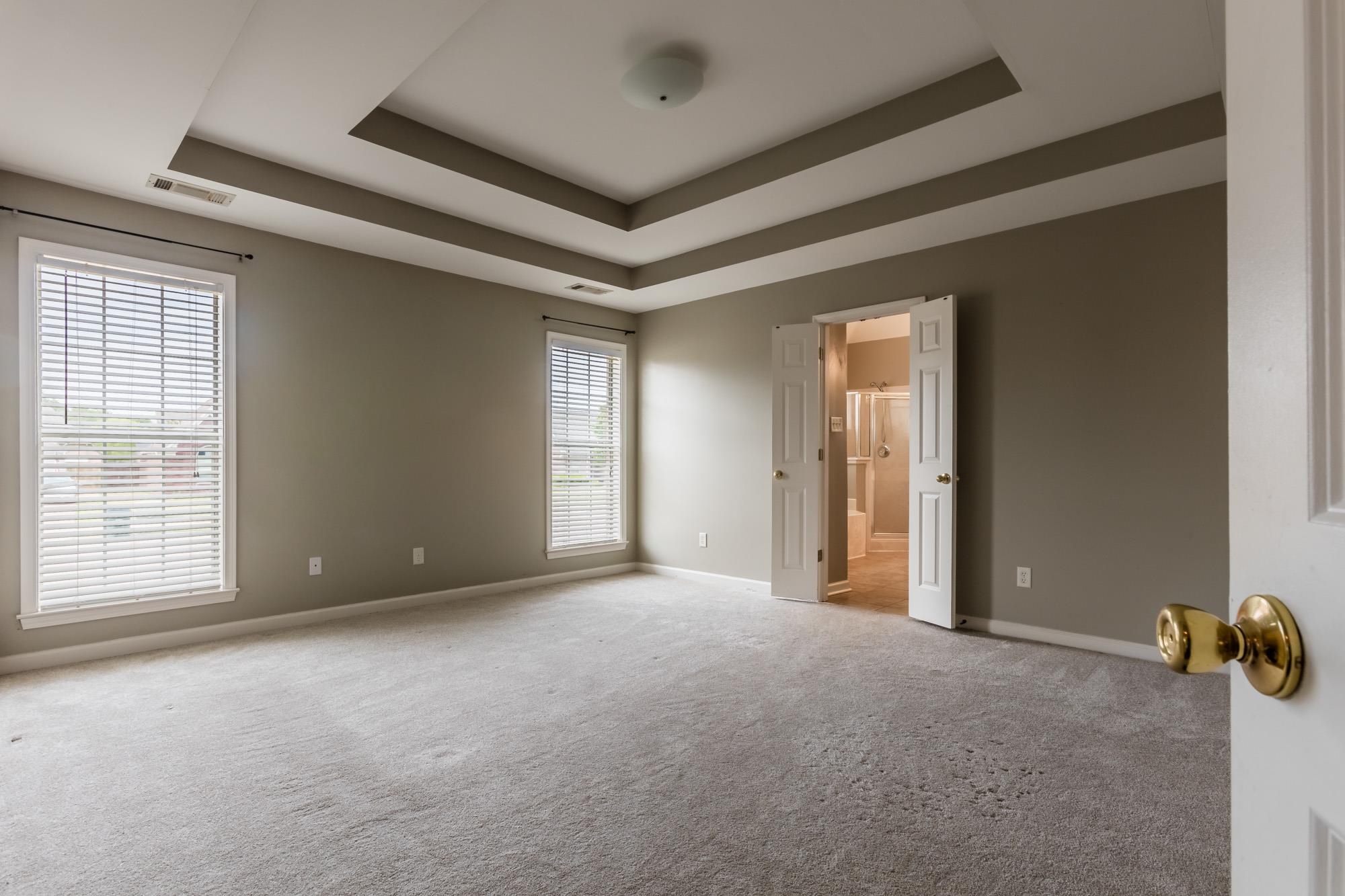 Empty bedroom with a tray ceiling, two blind-covered windows, and open doors to an ensuite bathroom.