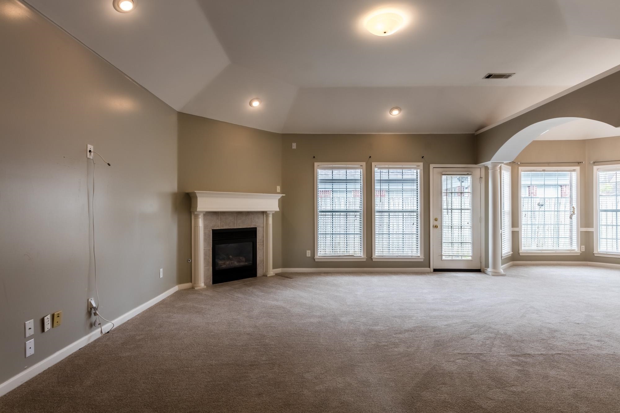Vacant living room with a tiled fireplace, vaulted ceiling, and wall of windows.