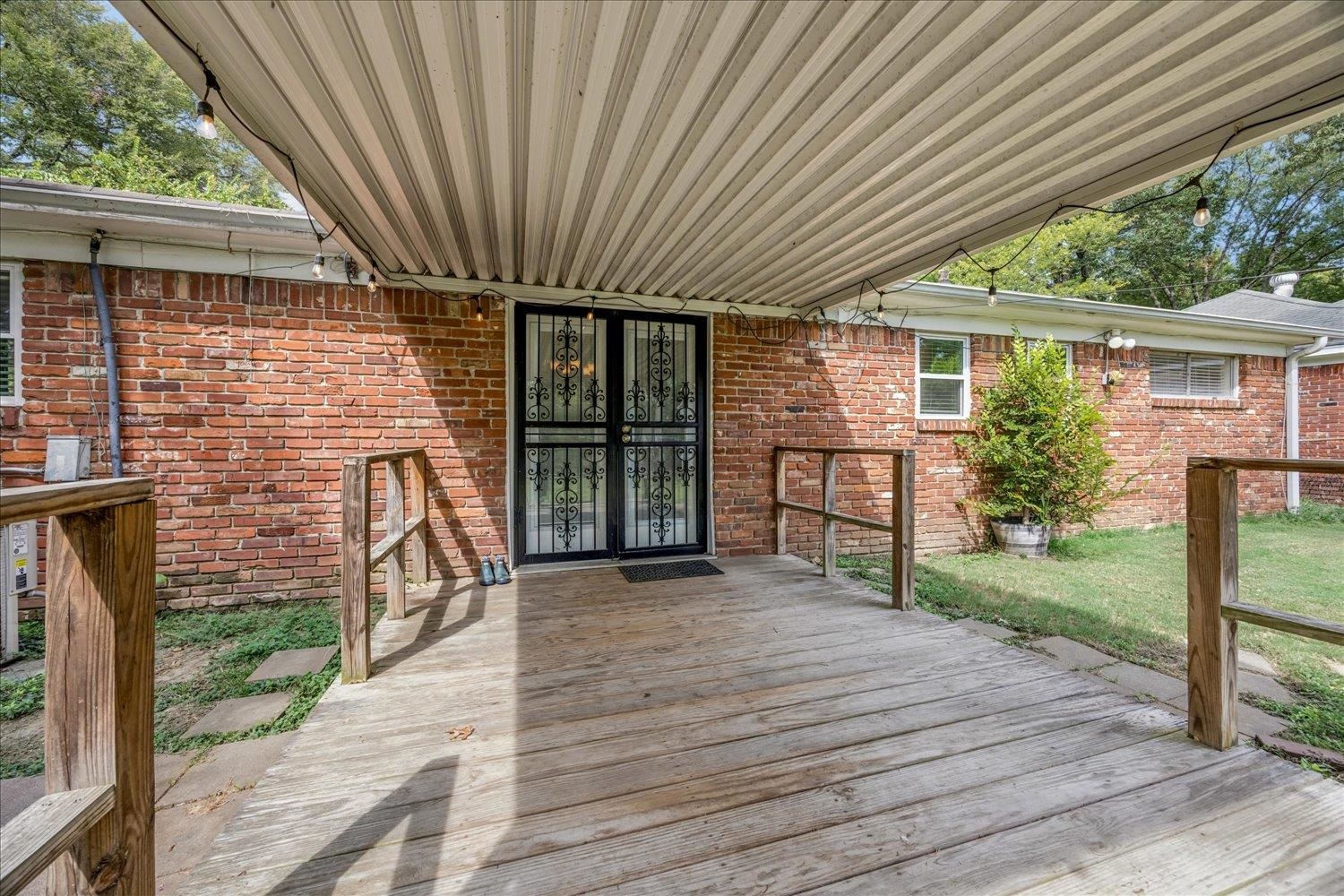 Covered wooden porch at a red brick house, with a black decorative security door and string lights.