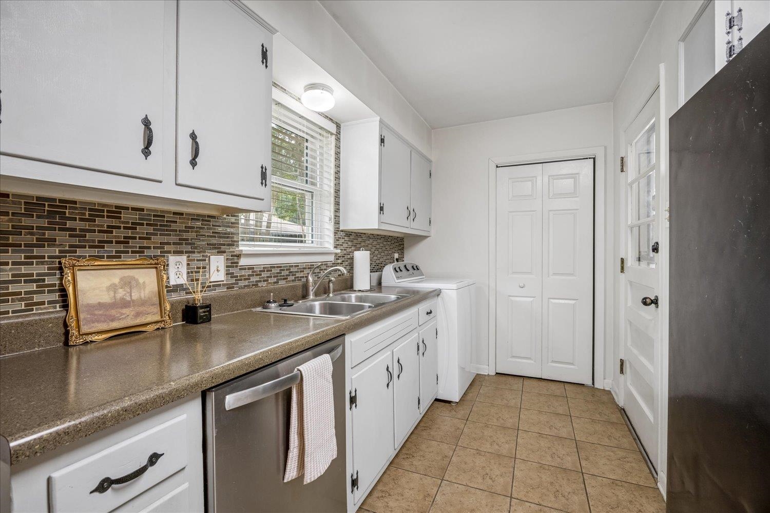 Kitchen with white cabinets, mosaic tile backsplash, stainless dishwasher, and sink beneath a window.