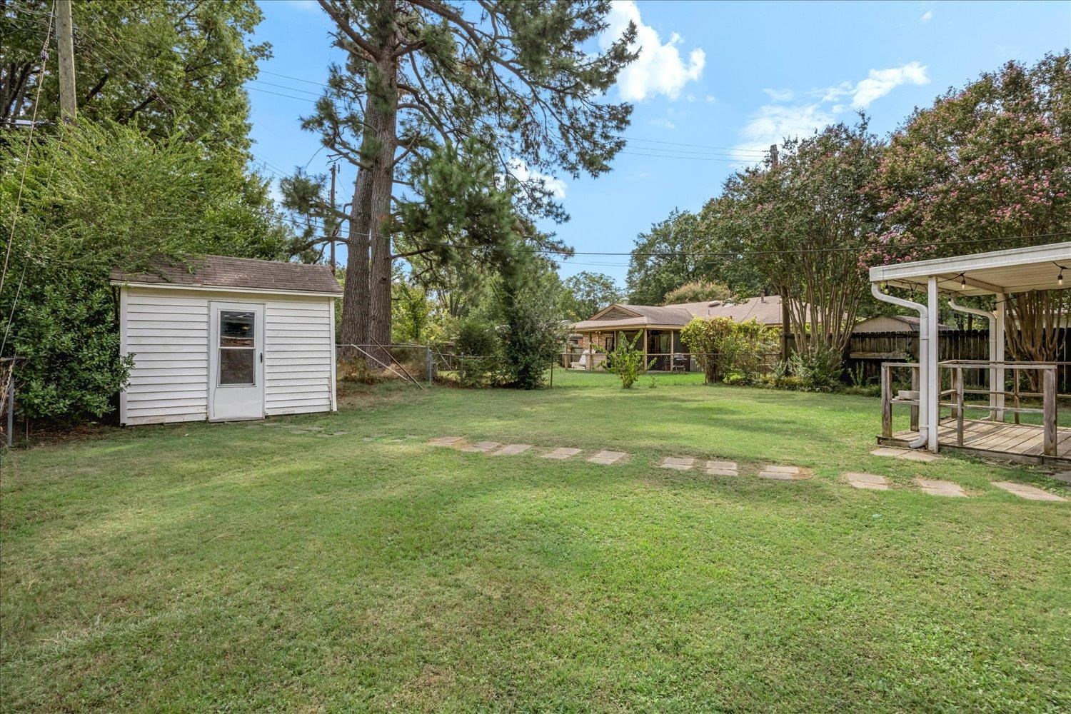 Backyard with a broad lawn, white storage shed, stepping stones, and covered deck beneath mature trees.