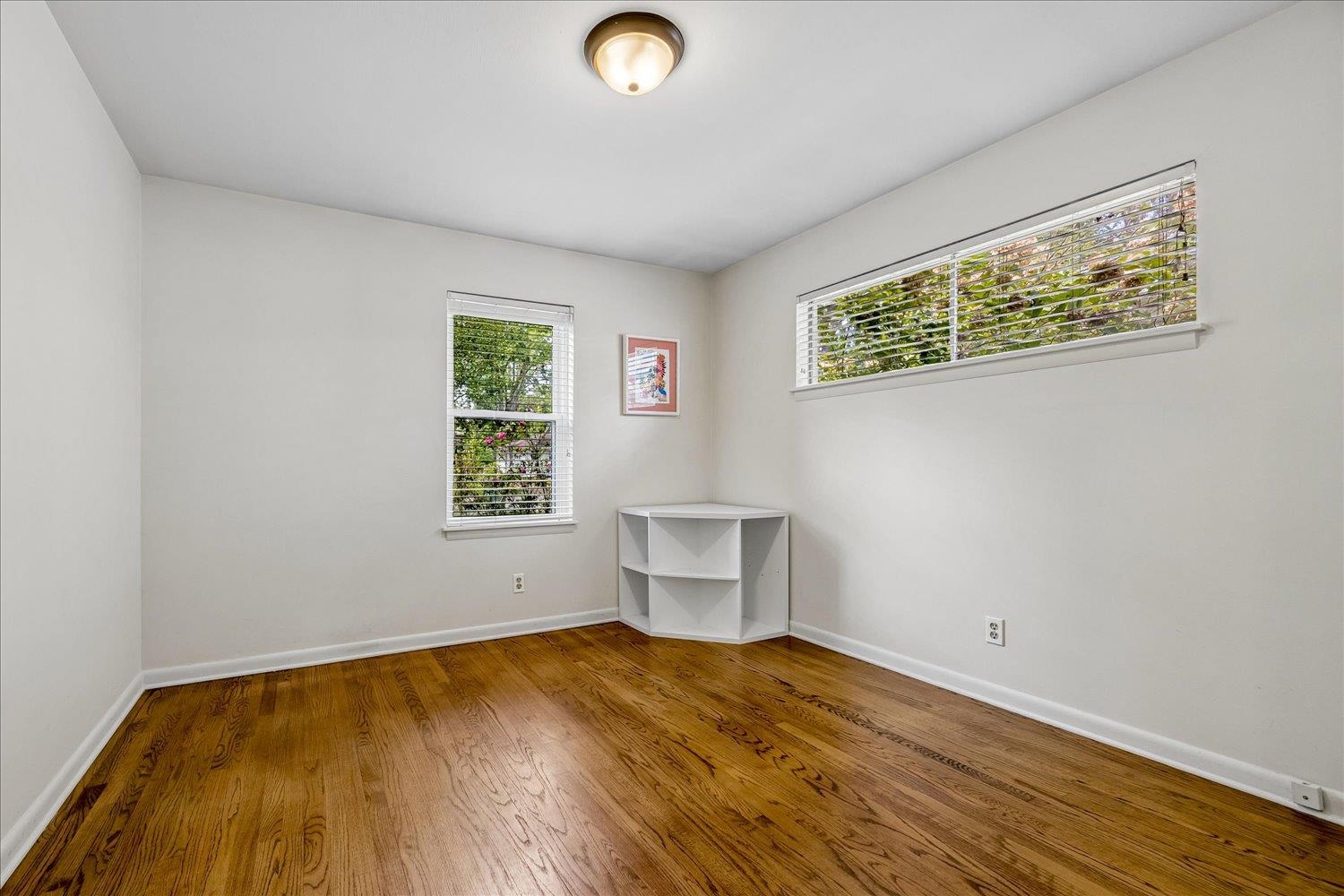 Empty room with polished hardwood floors, two windows with blinds, and a small corner shelf.
