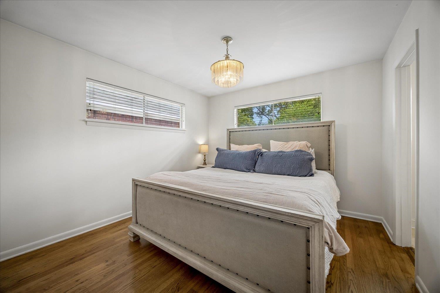 Bedroom with an upholstered bed, blue pillows, hardwood floors, and two high windows.
