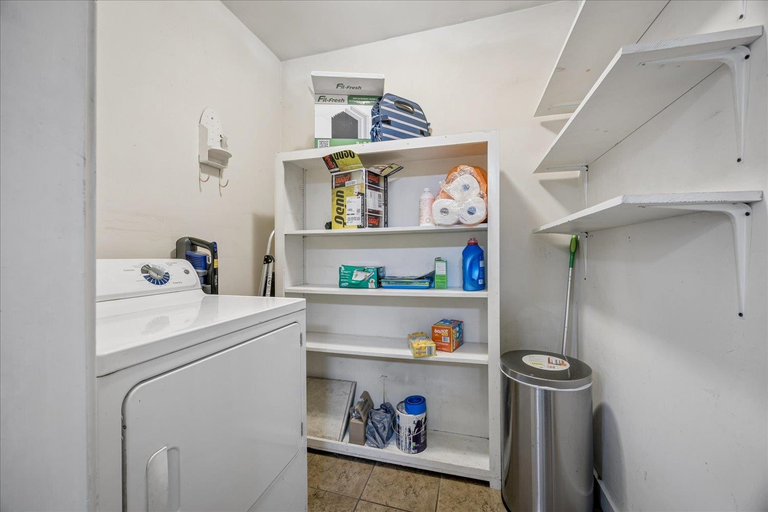 Laundry room with a white dryer, stocked storage shelves, and wall-mounted shelving.