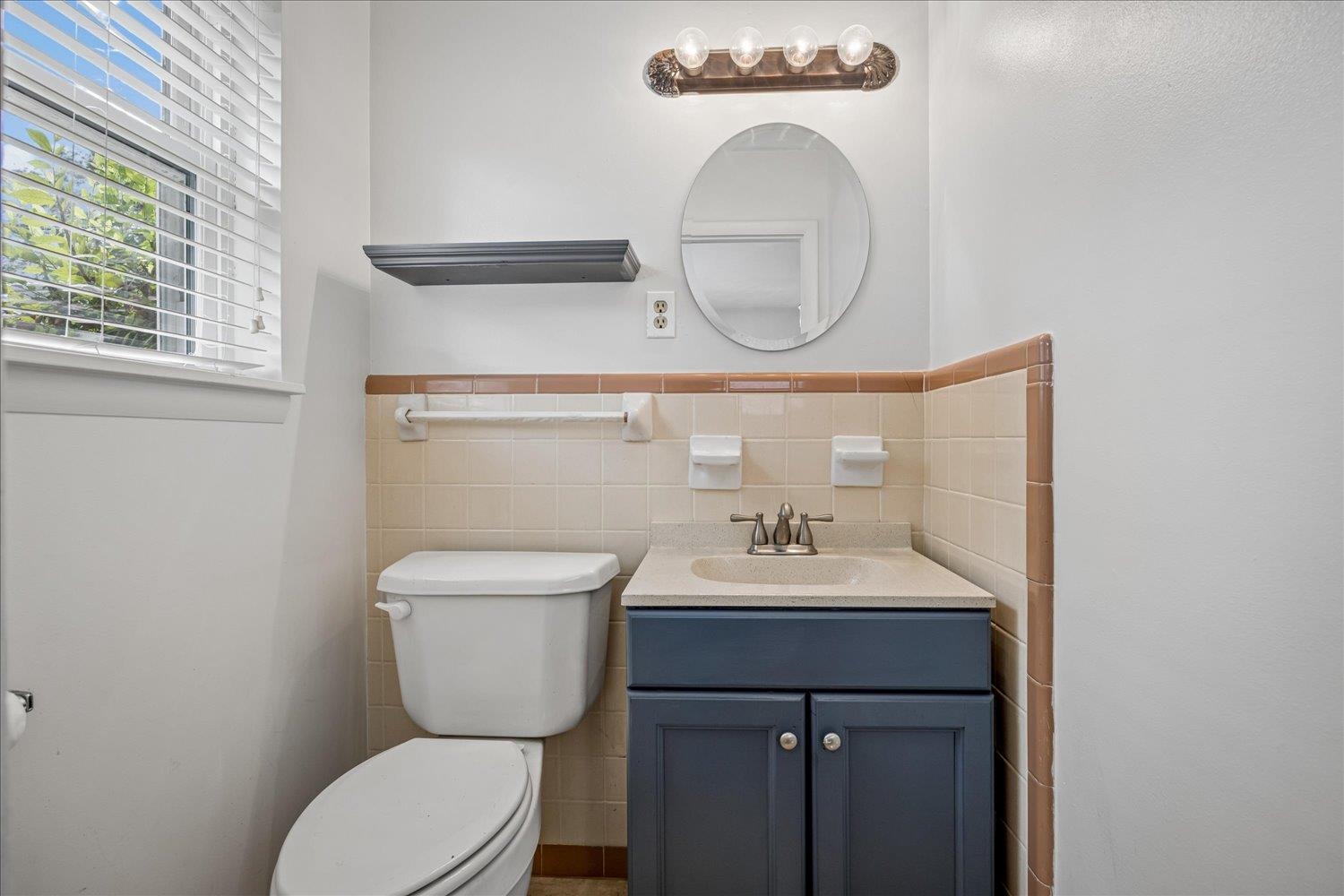 Bathroom with beige tile, a dark blue vanity, oval mirror, and window with white blinds.