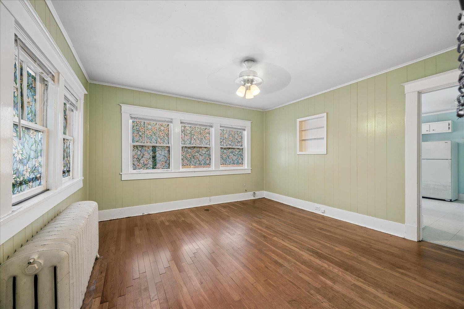 Empty room with hardwood floors, floral window coverings, and a doorway to the kitchen.