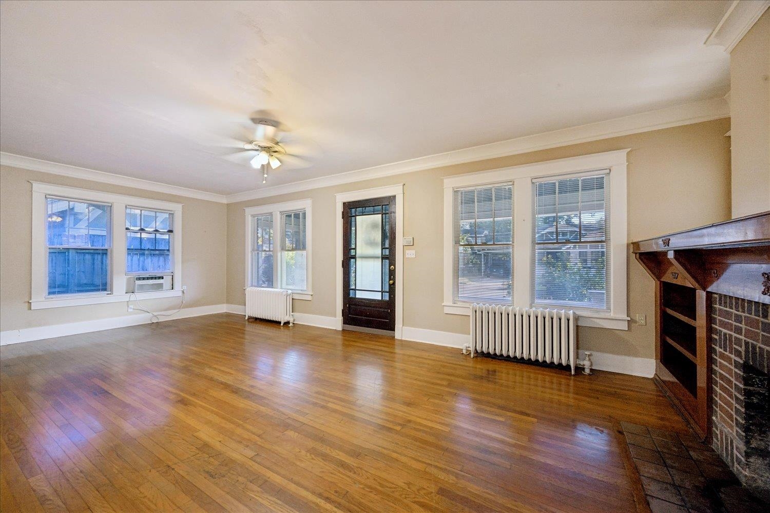 Empty living room with hardwood floors, tall windows, and a brick fireplace with a wooden mantel.