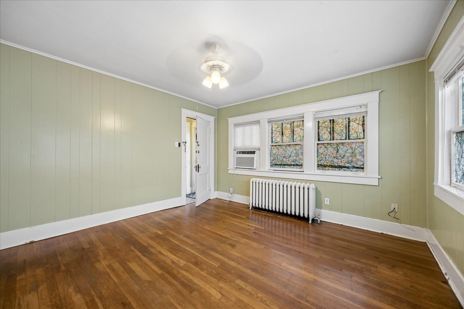 Empty room with hardwood floors, pale green paneled walls, floral stained-glass window panels, and a white radiator.