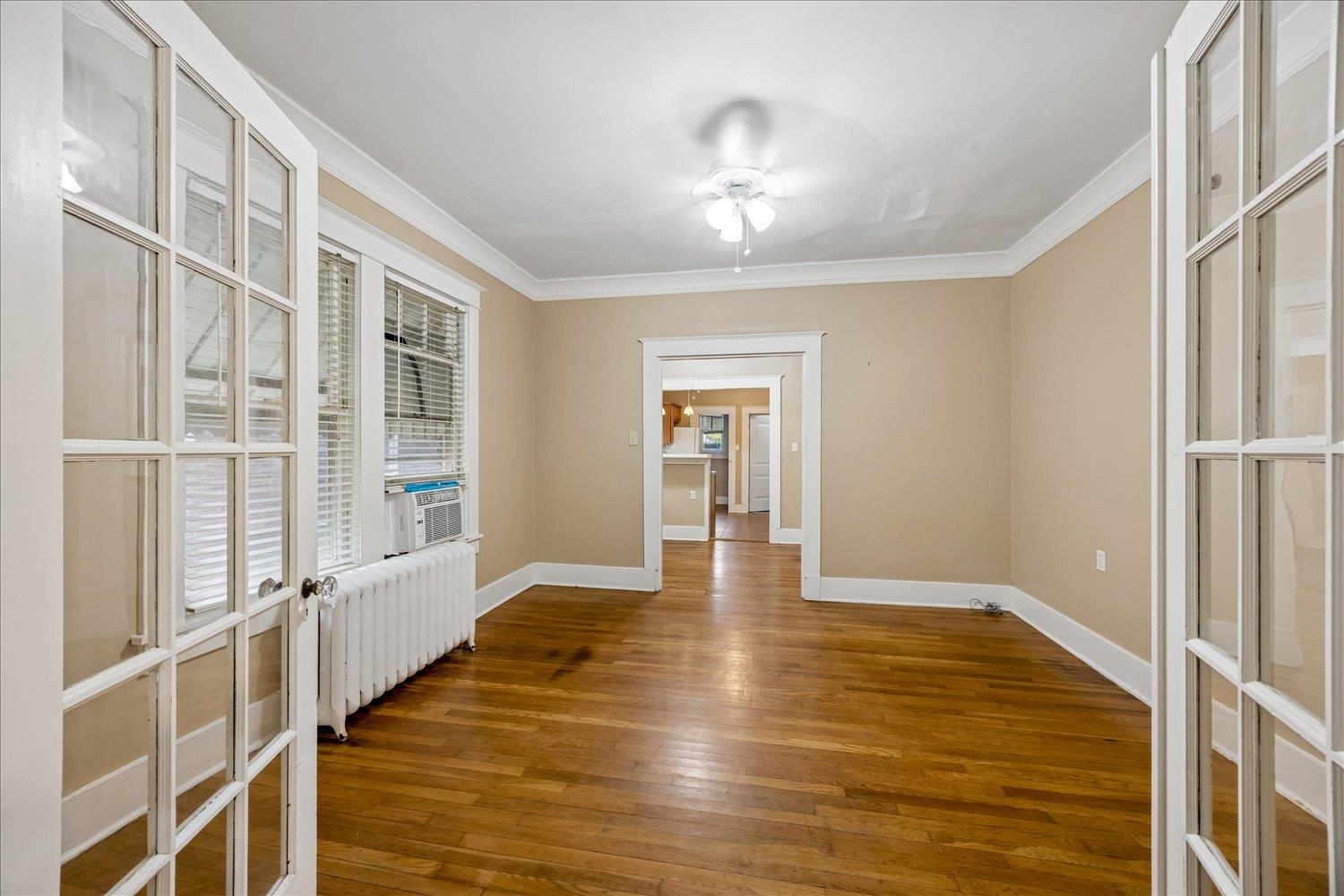 Empty room with hardwood floors, French doors, radiator, and doorway to the kitchen.