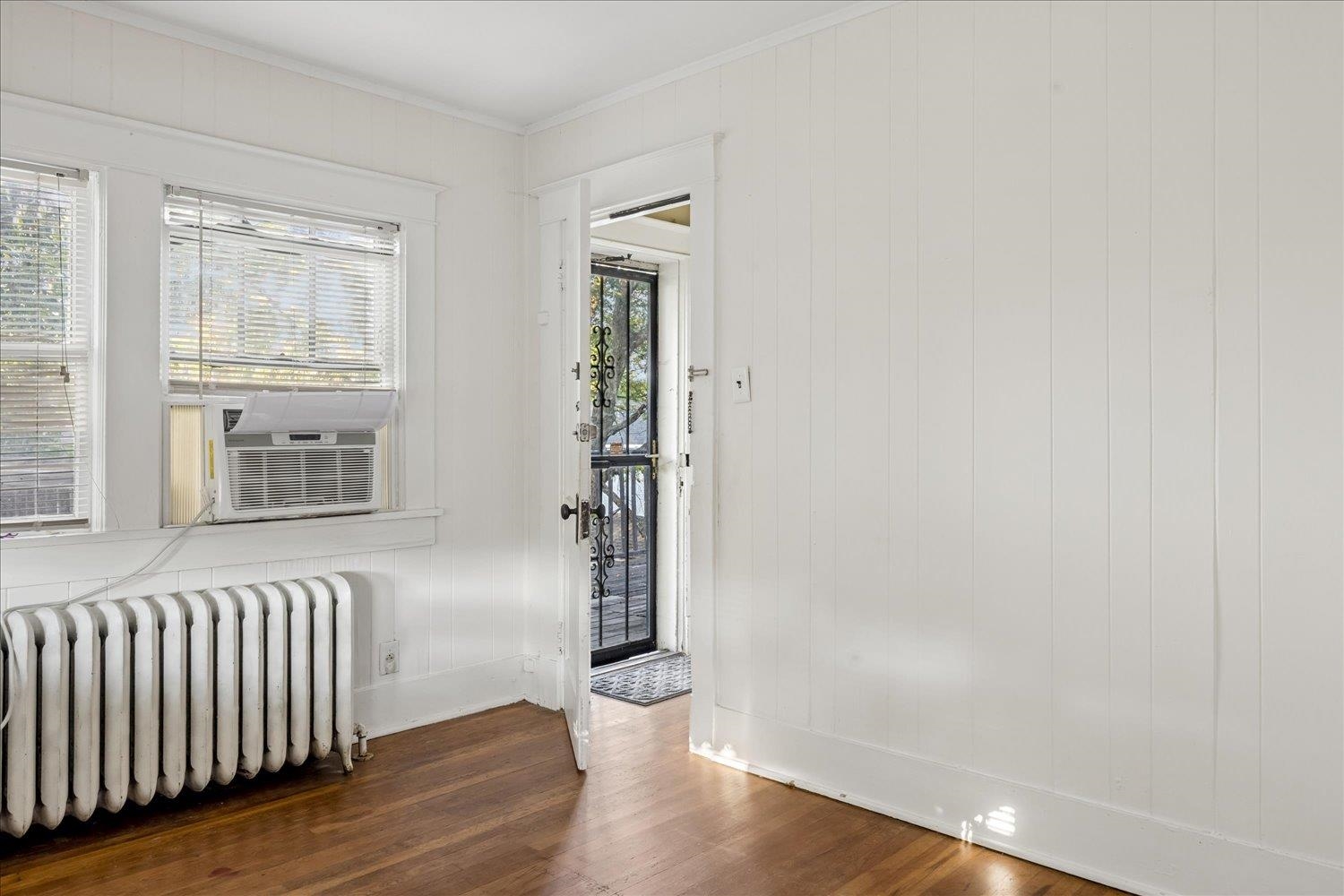 Empty room with hardwood floors, a cast-iron radiator, window air conditioner, and open doorway to an exterior landing.