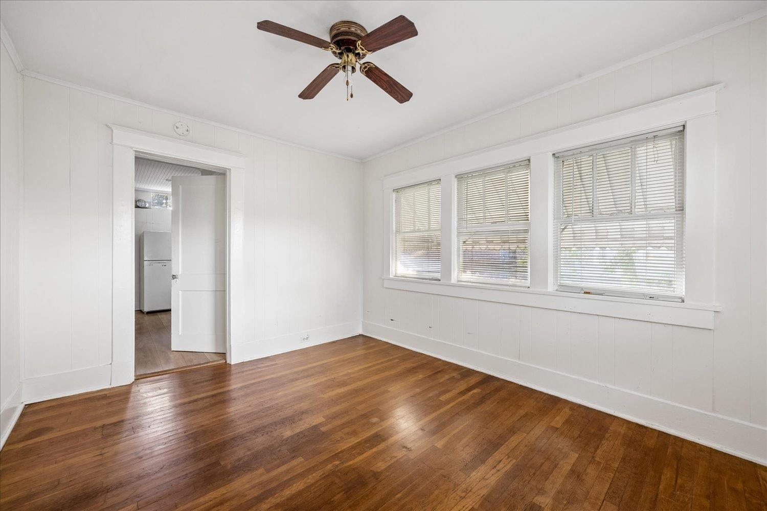 Empty room with hardwood floors, three windows, and a ceiling fan.