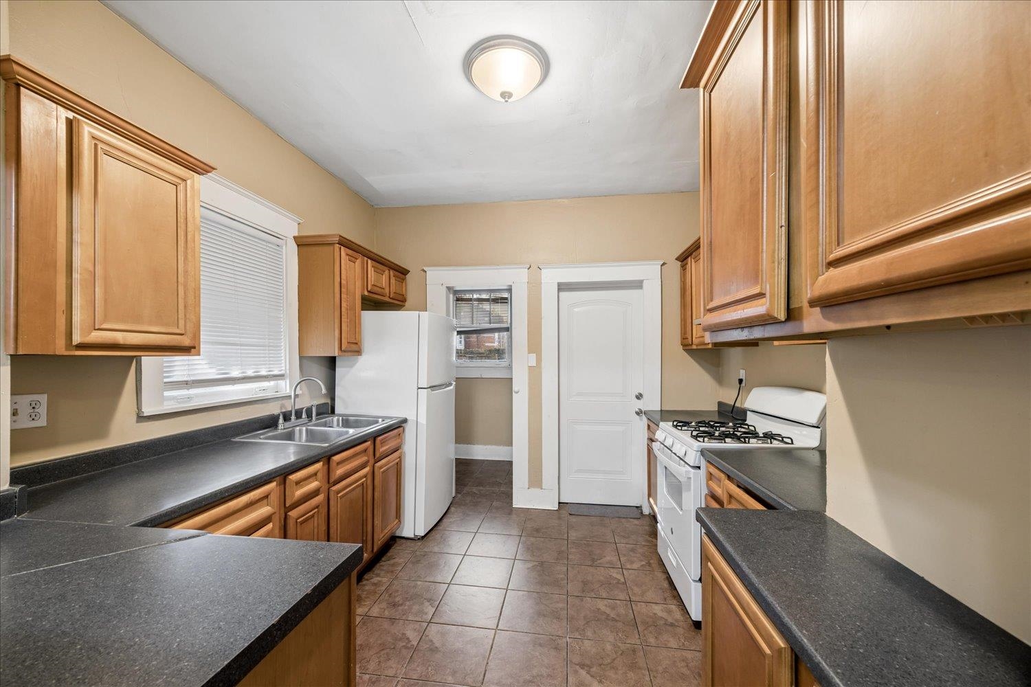 Kitchen with wood cabinets, dark countertops, a double sink, and white appliances.