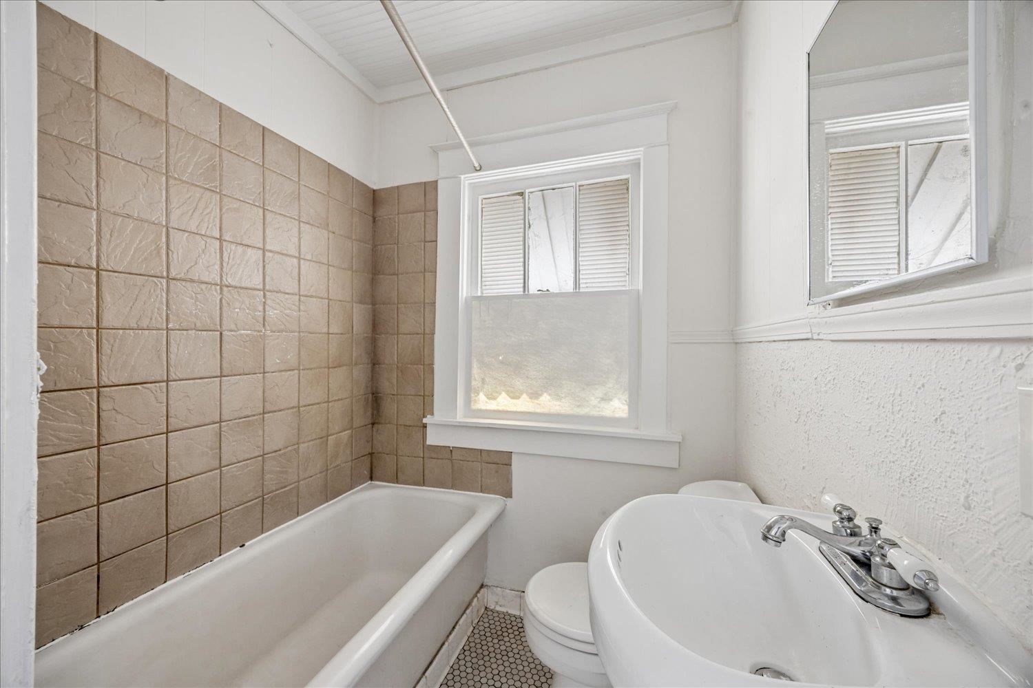 Bathroom with a beige tiled tub surround, pedestal sink, and window with a translucent shade.
