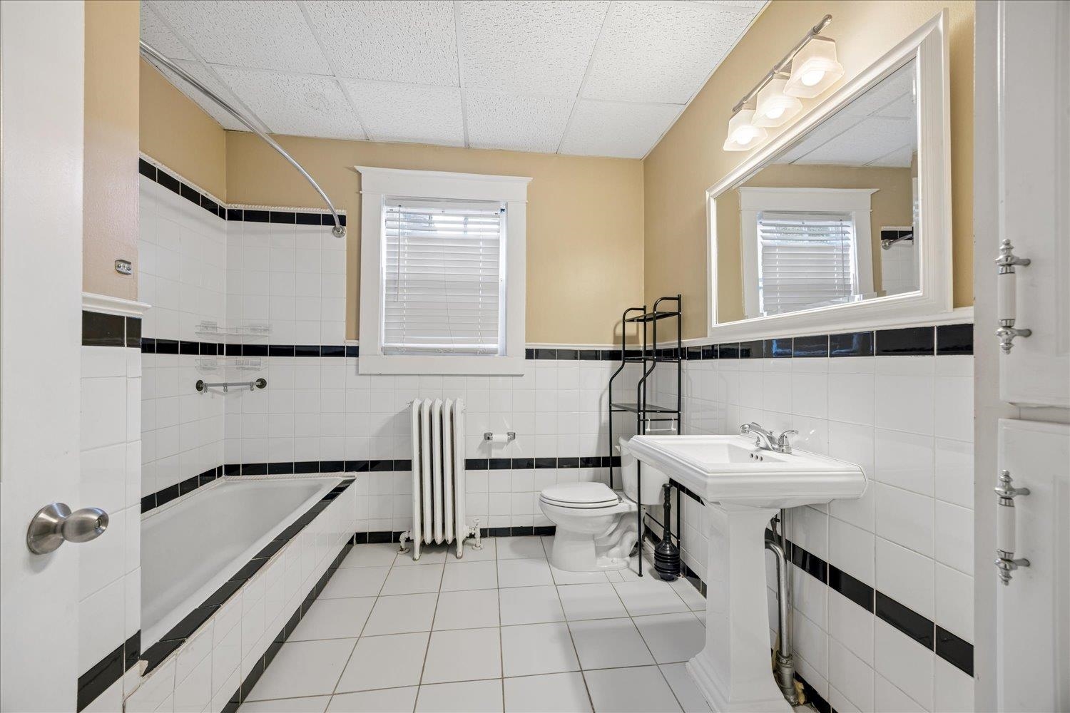 Bathroom with a tiled bathtub, pedestal sink, radiator, and black tile accents.