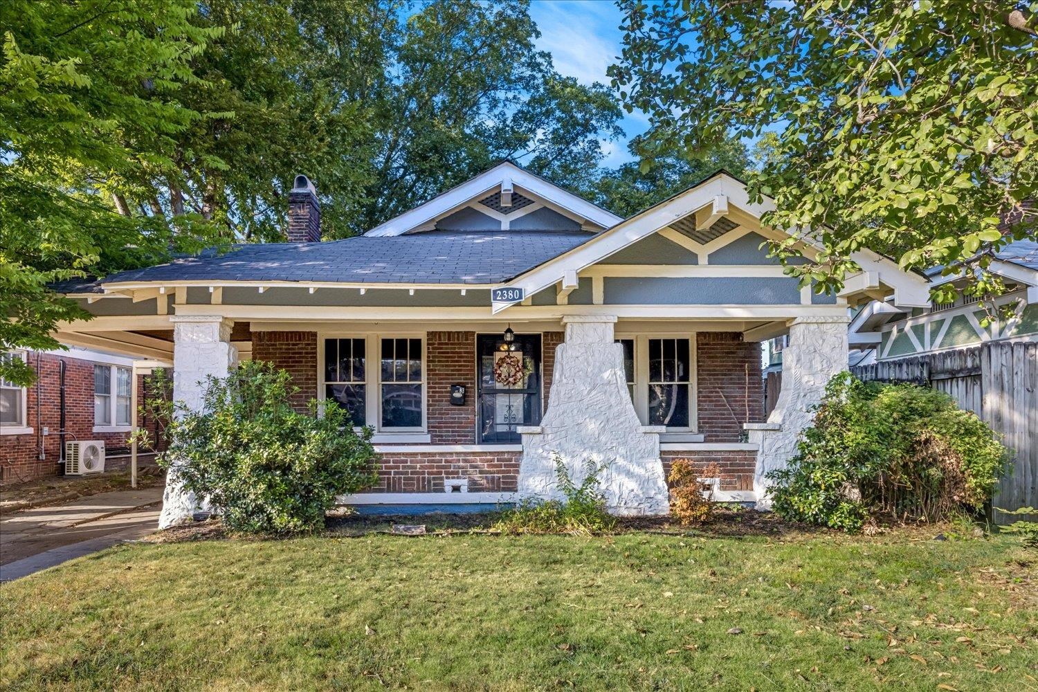 Brick bungalow with a broad covered porch, white stone columns, and mature trees.
