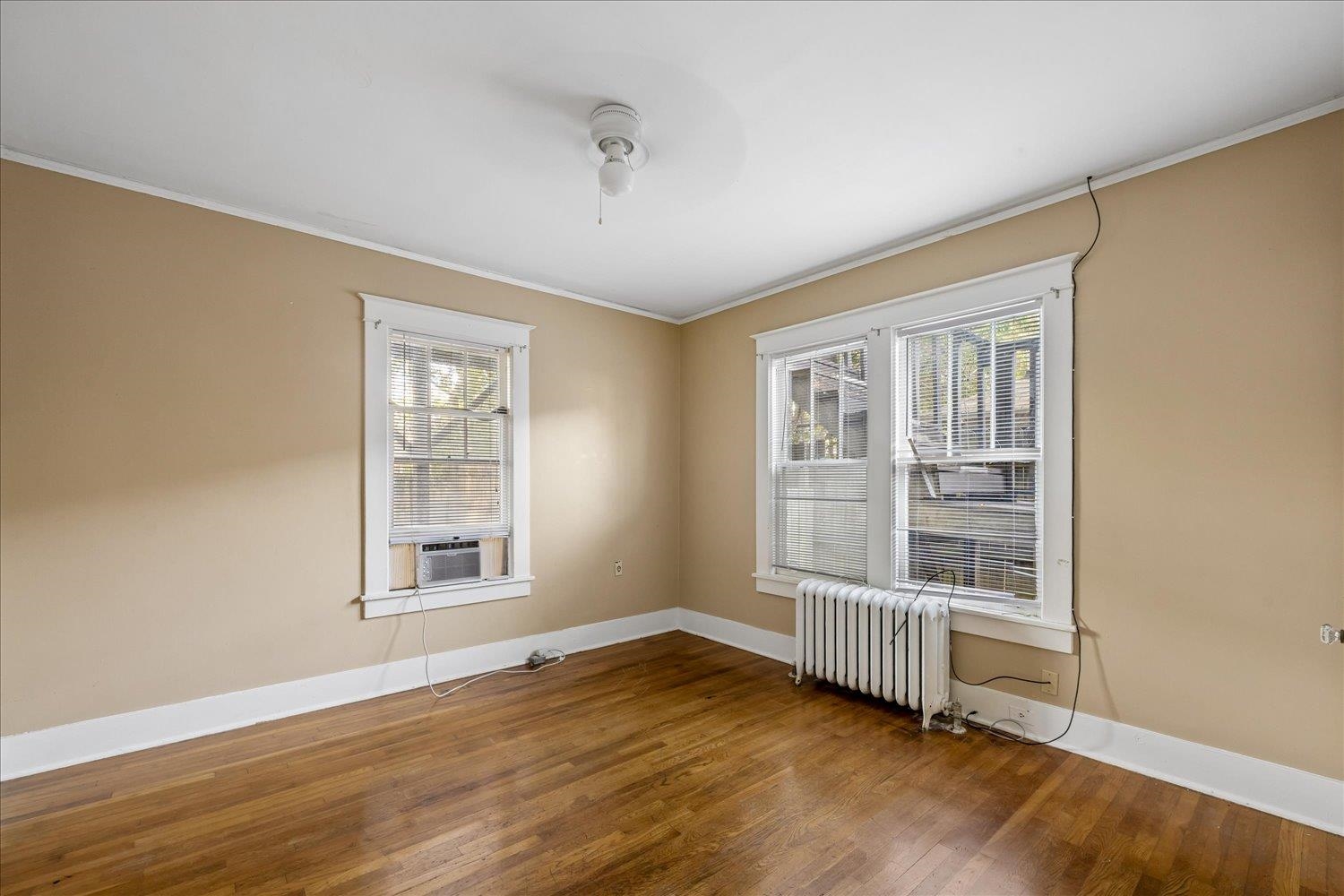 Empty room with hardwood floors, tan walls, two windows, a window air conditioner, and a radiator.