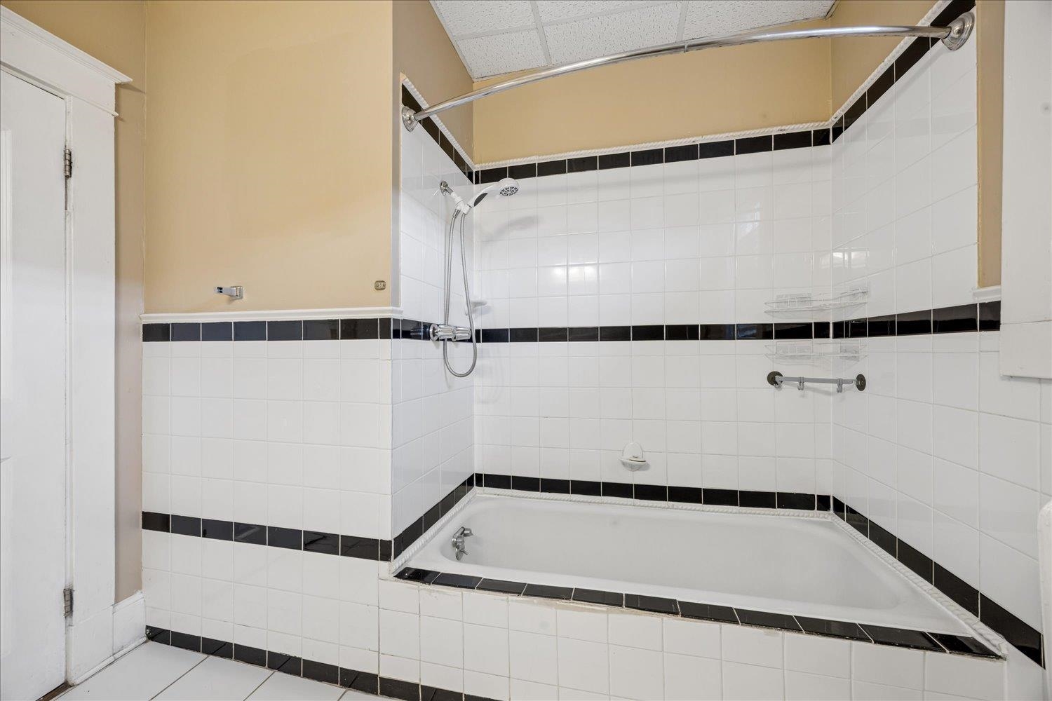 Bathroom with a built-in tub, white tile surround, black accent bands, and handheld showerhead.