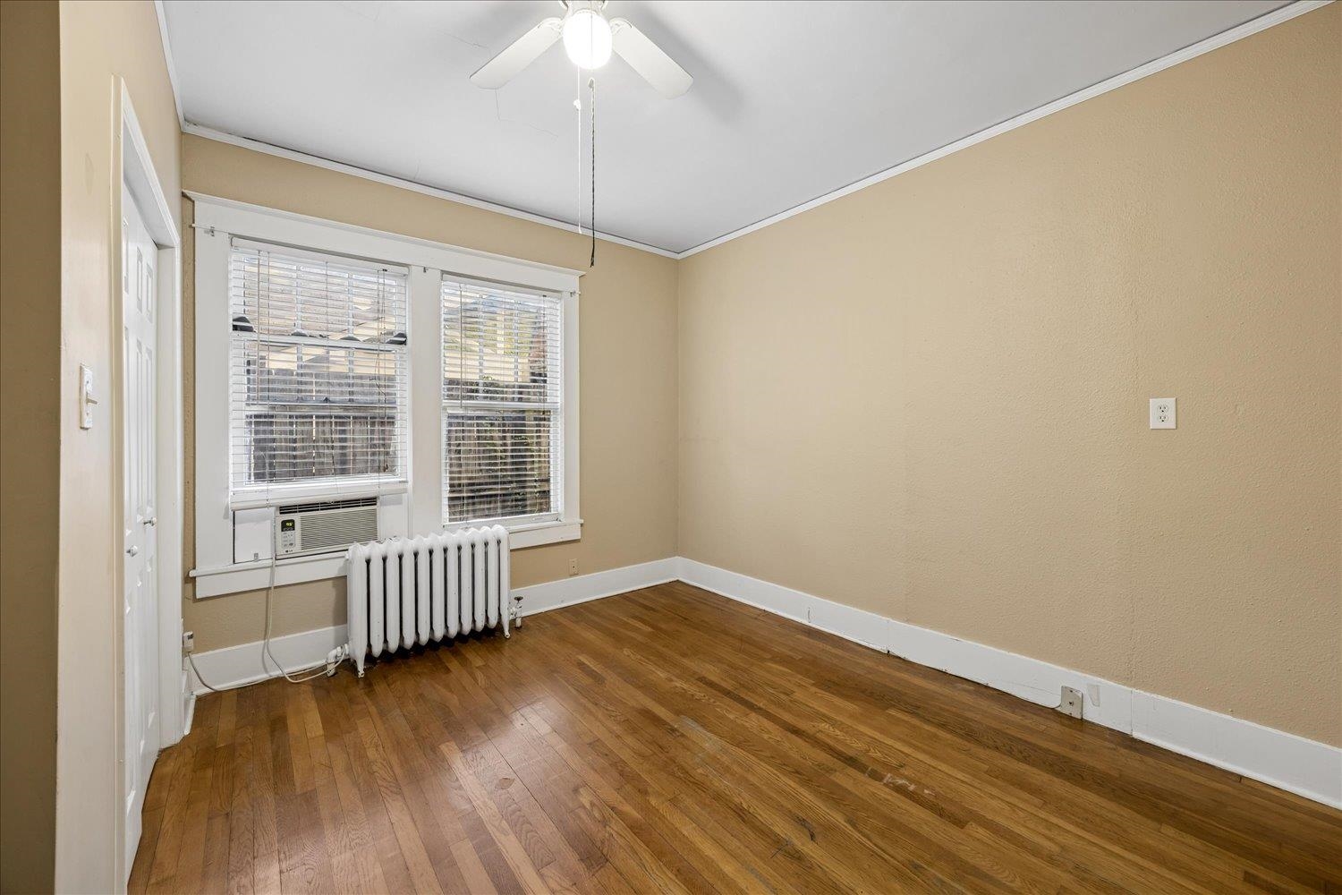 Empty room with hardwood floors, two blinded windows, a radiator, and a ceiling fan.