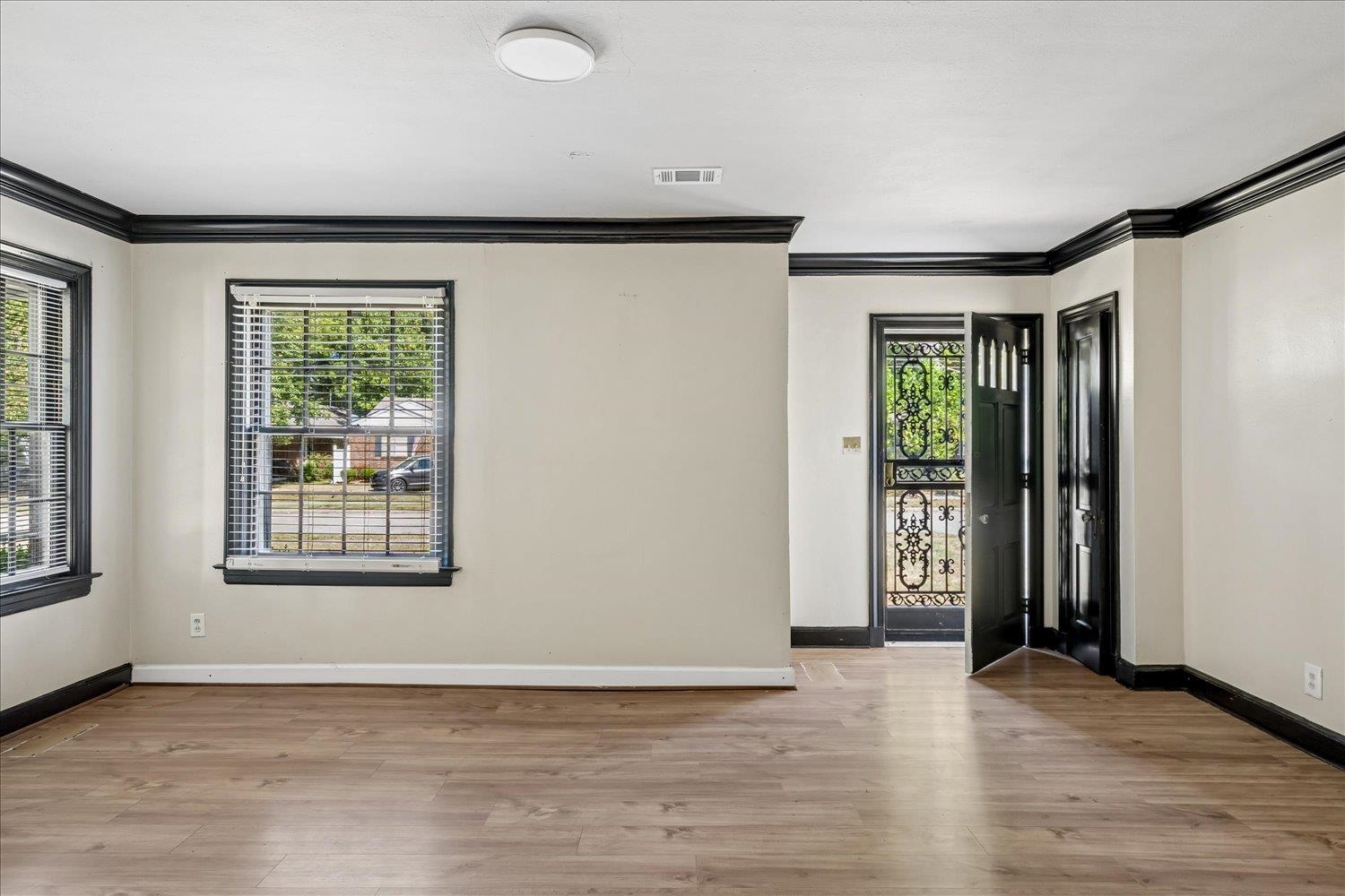 Empty front room with light wood floors, black trim, and an open entry door with decorative metalwork.
