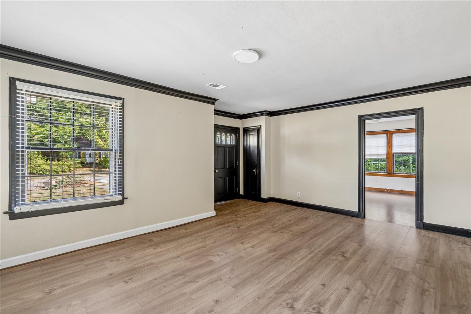 Empty living room with light wood floors, dark trim, and a large window with blinds.