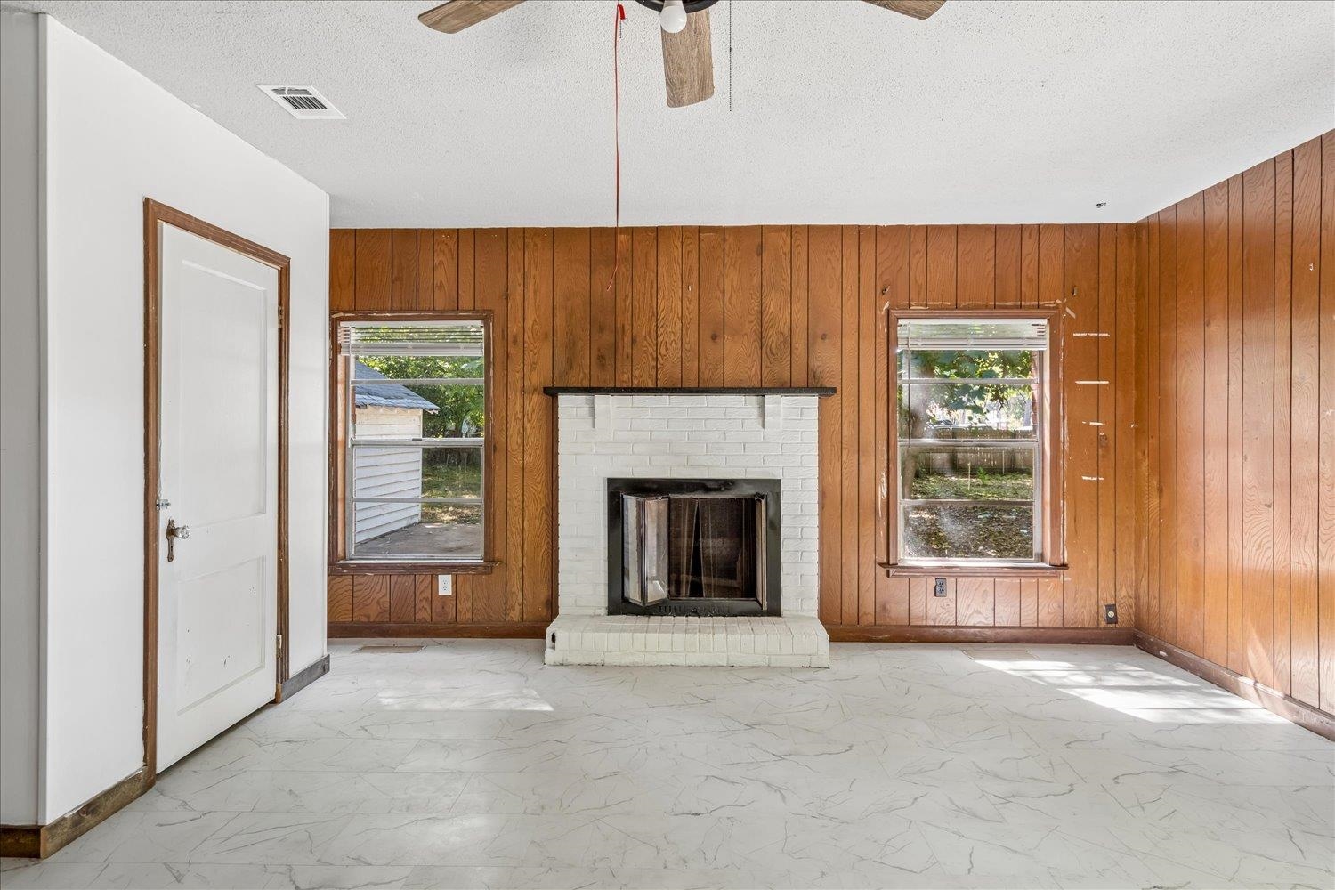 Empty living room with wood-paneled walls, a white brick fireplace, and marble-patterned flooring.