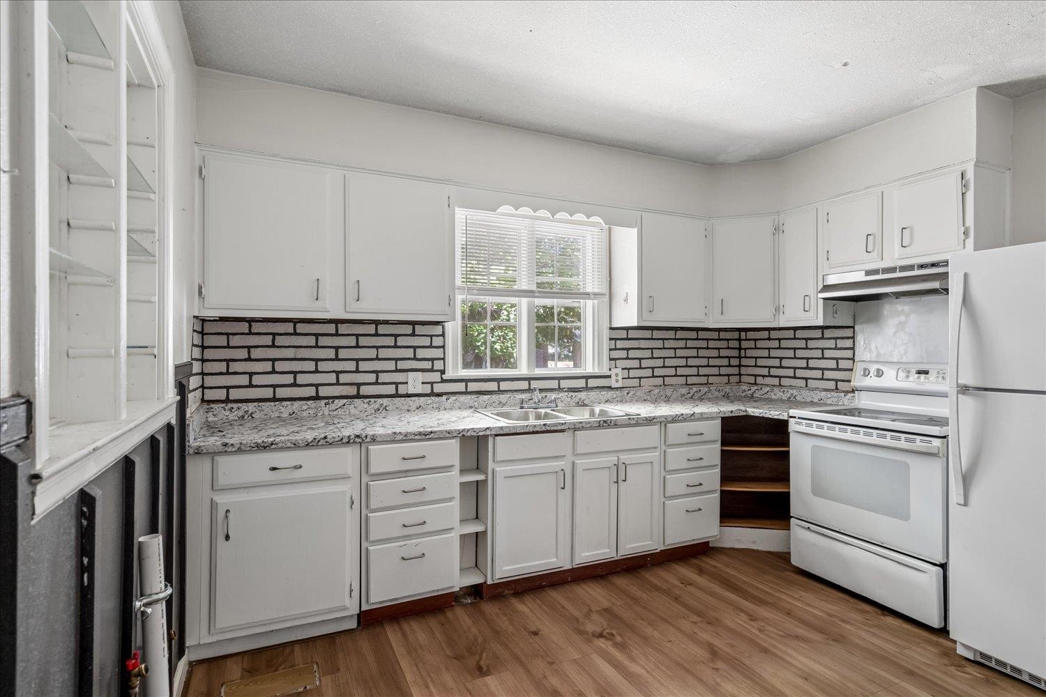 Kitchen with white cabinets, brick backsplash, double sink beneath a window, and wood-look flooring.
