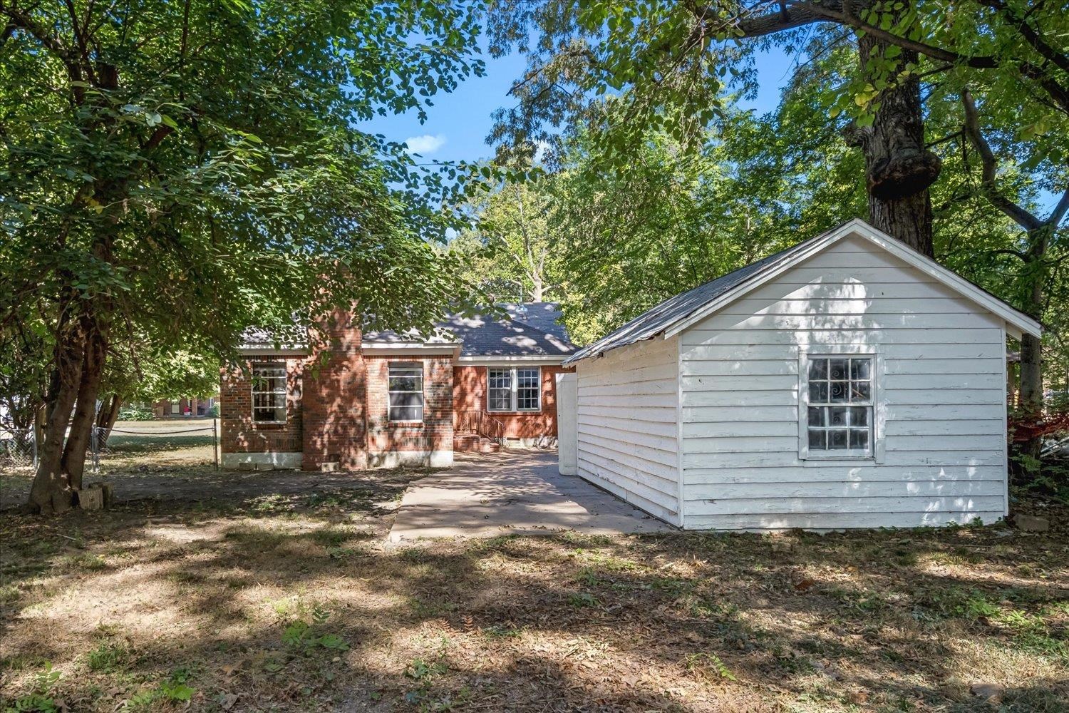 Rear of a brick home and white detached shed beneath mature trees.